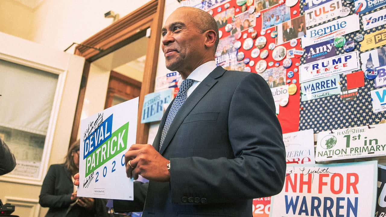 穿西裝的男子被政治海報包圍，舉著Deval Patrick 2020競選標語牌。