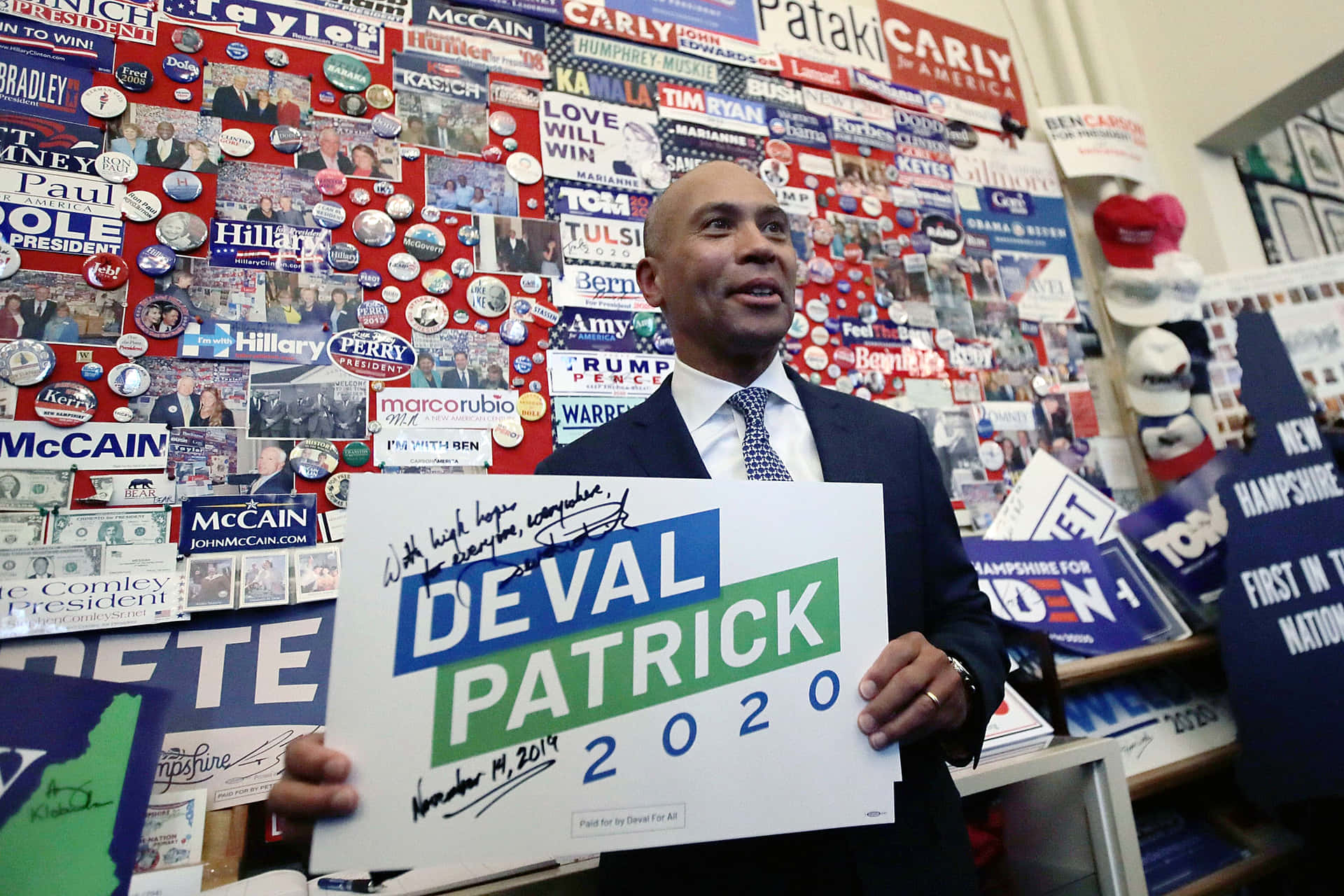男子舉起 Deval Patrick 2020 競選標語牌，周圍環繞著政治紀念品。