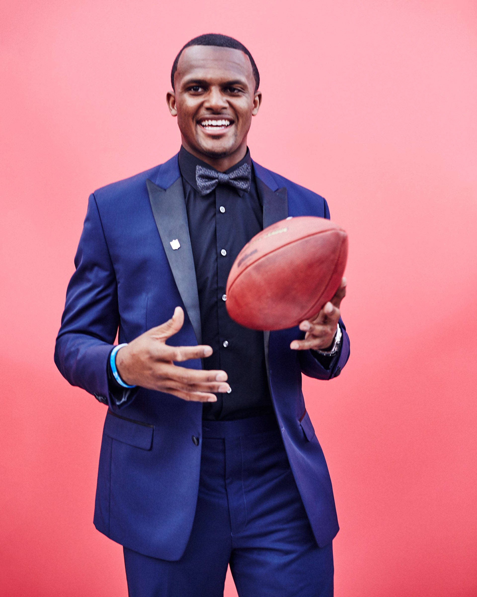德肖恩·沃森2017年NFL選秀 壁紙 — 一位笑容滿面的男士身穿筆挺藍色西裝，在充滿活力的粉紅色攝影棚背景前抱著一個足球。