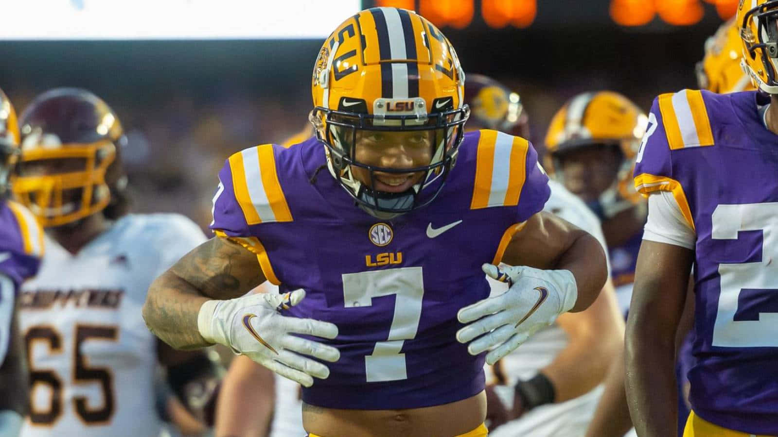 德里克·斯廷格利 Jr. LSU 騎士隊超級明星 壁紙 — 一位身穿紫色和金色制服的路易斯安那州立大學足球員在體育場燈光下比賽時微笑，雙手放在胸前。