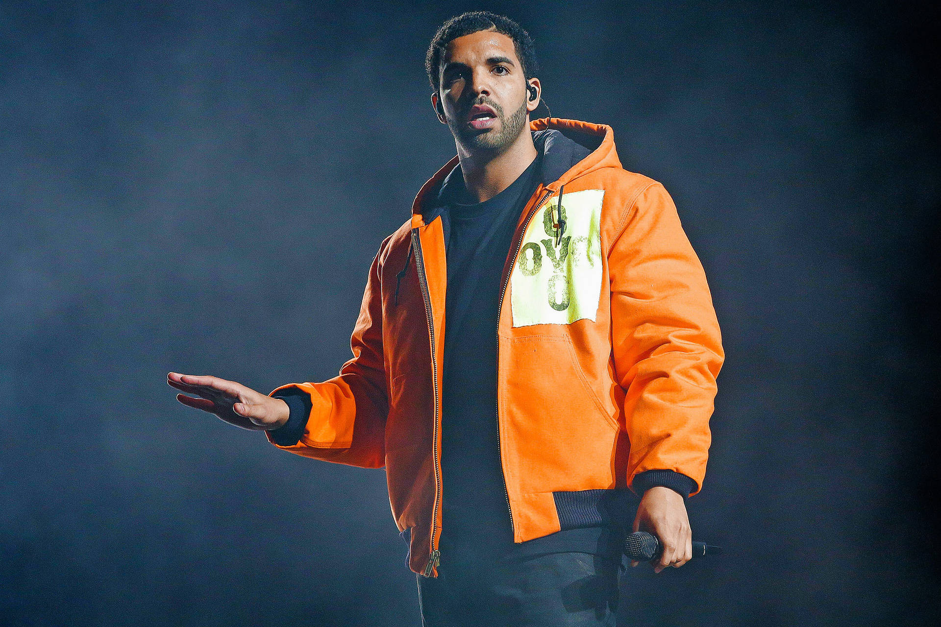Drake 在舞台上表演，身穿橙色外套，手持麥克風。