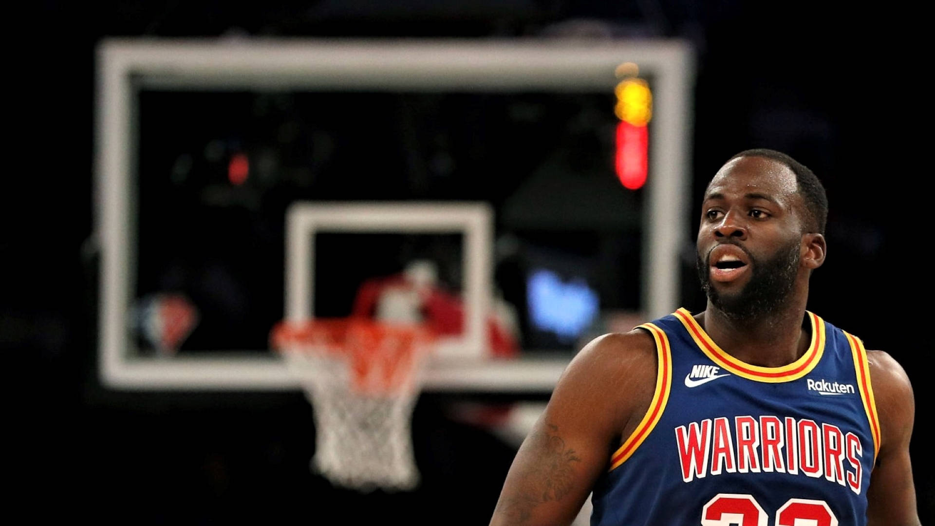 德雷蒙德·格林 NBA 球員 壁紙 — 一位身穿勇士隊球衣的專注籃球運動員在比賽中於場館燈光下凝視，身後有一個模糊的籃框。