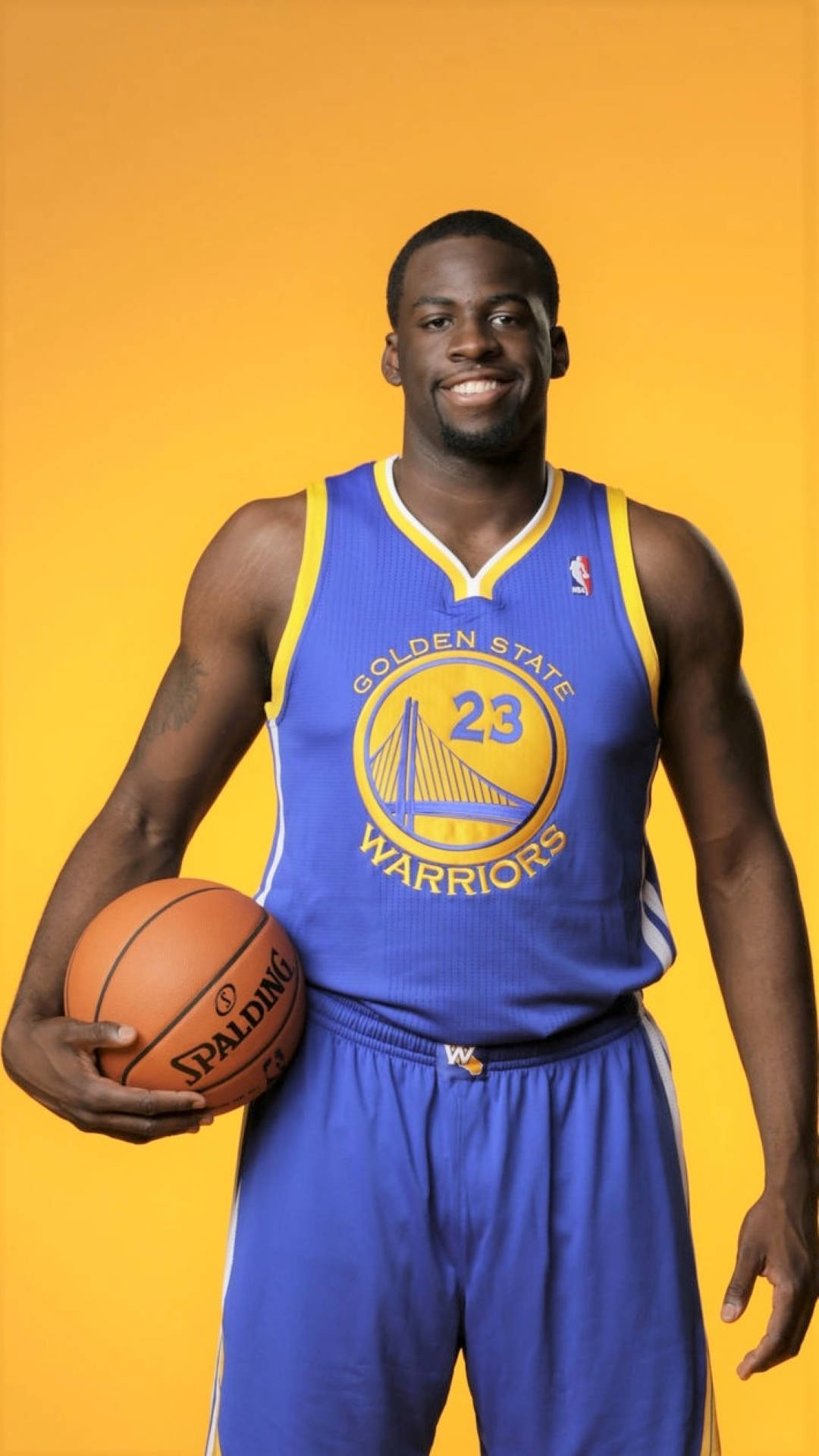 德雷蒙德·格林2012年NBA新秀 壁紙 — 一位微笑的籃球運動員身穿金州勇士隊球衣，在純黃色背景前拿著一個Spalding籃球。