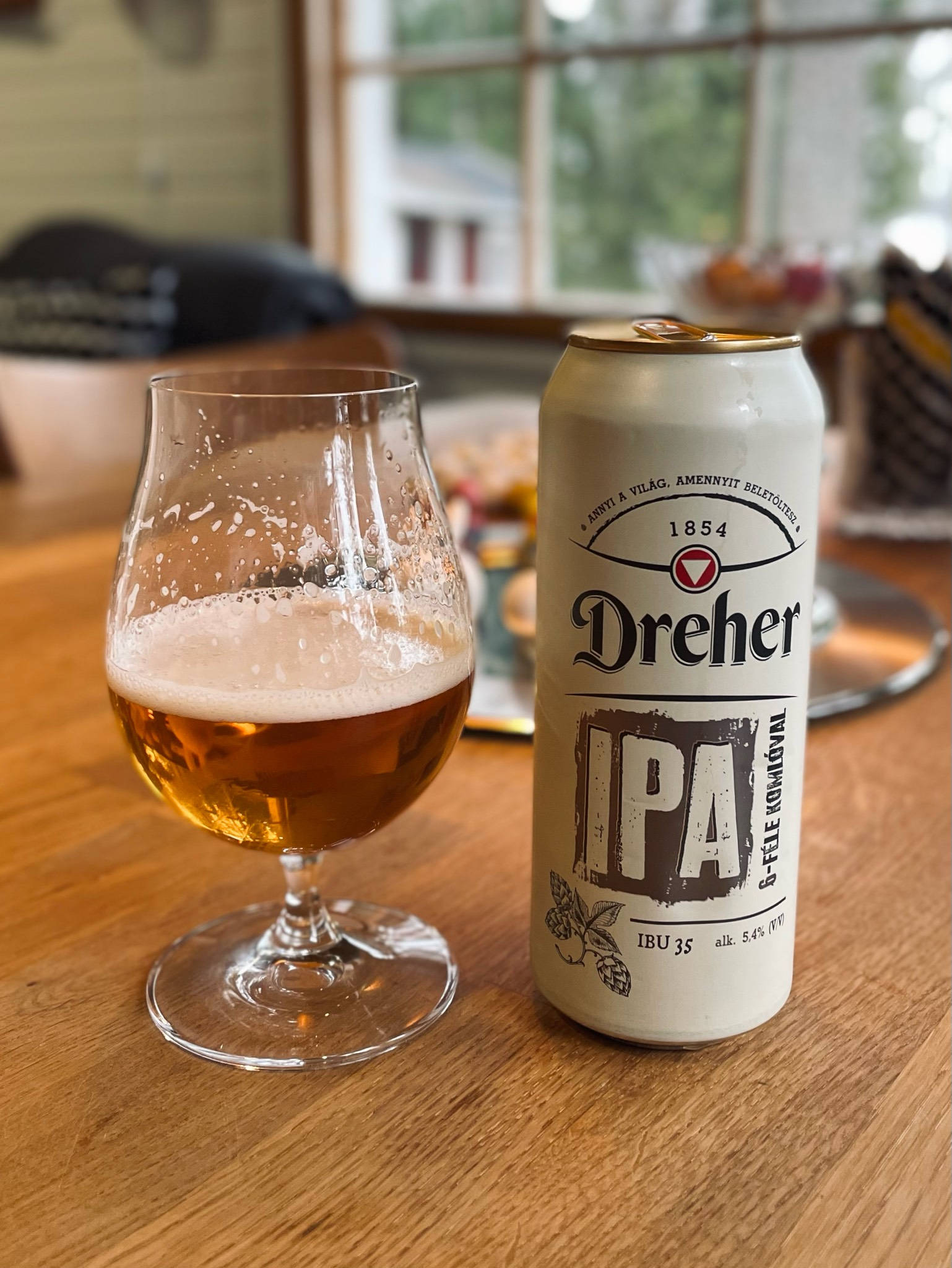 德雷赫 IPA 啤酒 壁紙 — 一杯泡沫豐富的金色IPA啤酒放在鬱金香杯中，旁邊放著它的罐頭，在窗邊溫暖的木桌上。