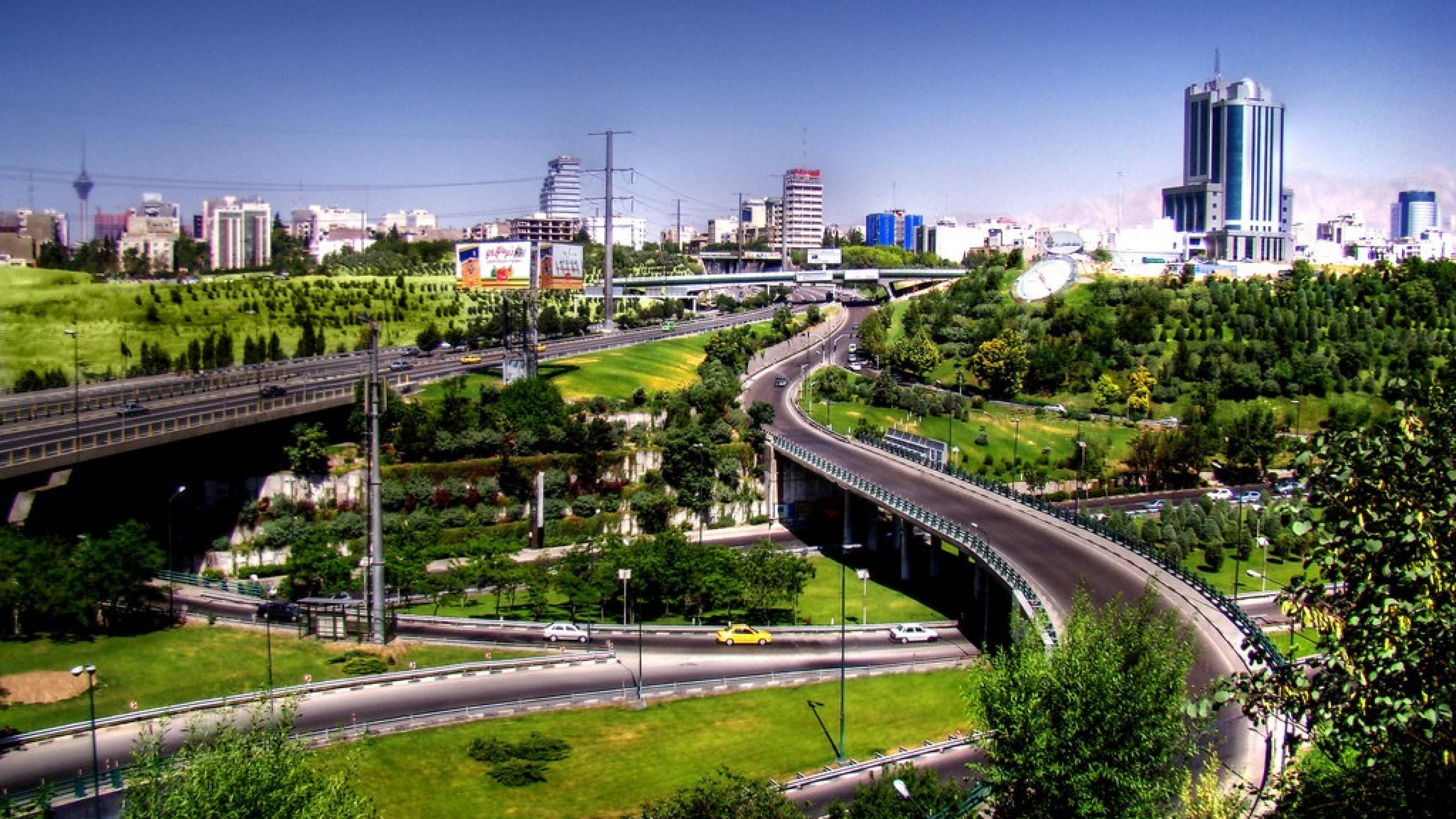 德黑蘭城市快速道路 壁紙 — 廣闊的城市景觀，蜿蜒的高速公路穿過鬱鬱蔥蔥的綠色公園和現代摩天大樓，在清澈的藍天下。