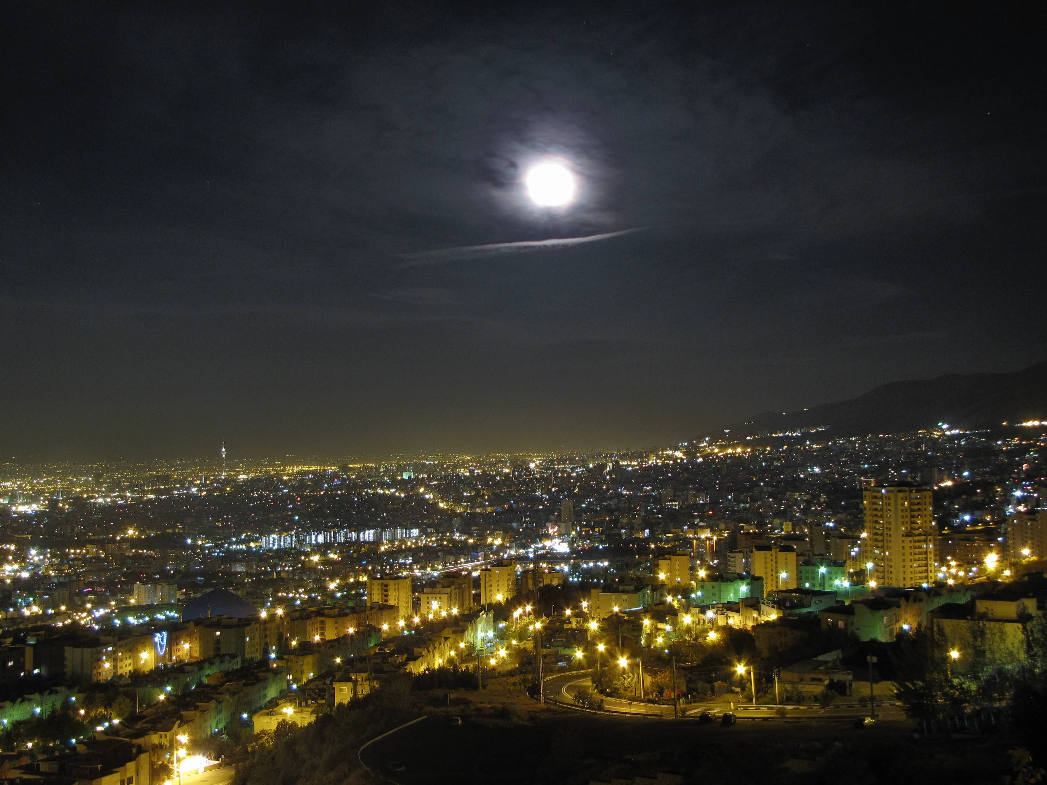 德黑蘭滿月夜 壁紙 — 一輪明亮的滿月懸掛在夜晚廣闊的城市景觀上空，其光芒照亮了下方無數的路燈和建築物。