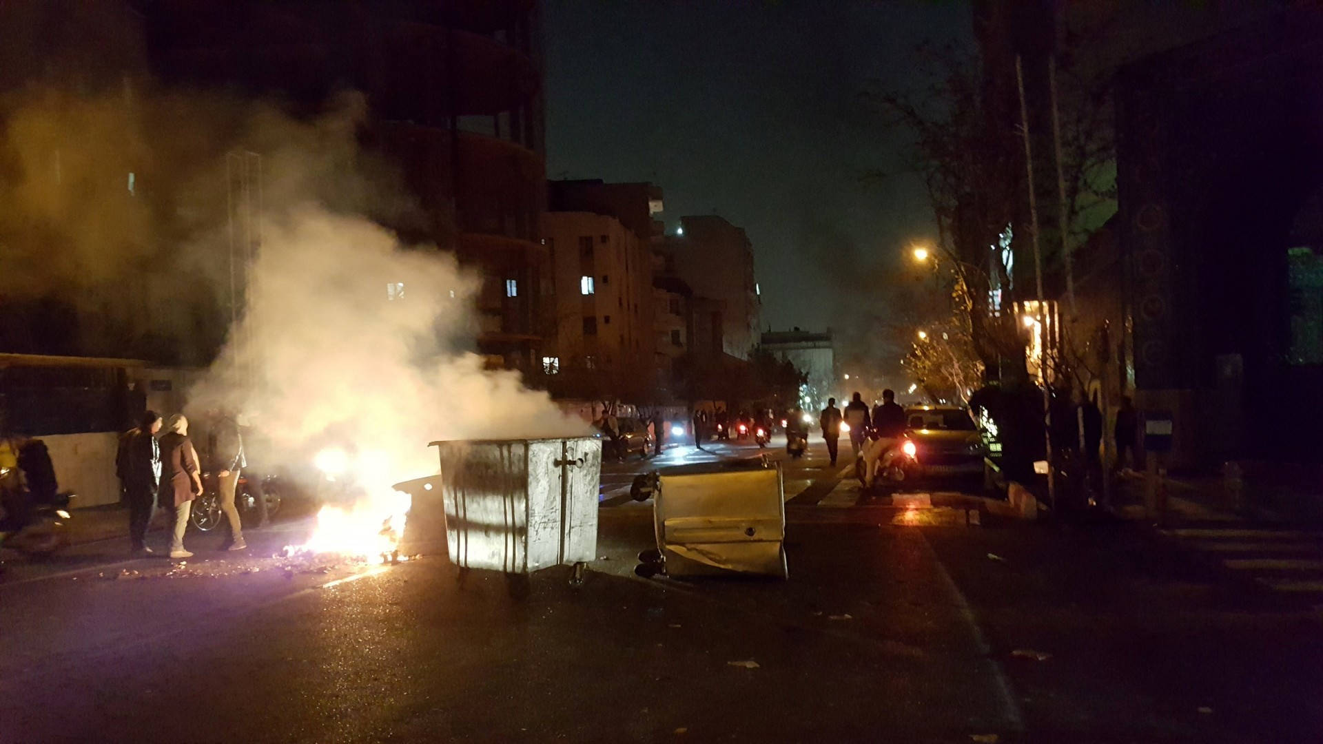 德黑蘭火災抗議 壁紙 — 一個垃圾箱在夜晚黑暗的城市街道上猛烈燃燒，發出橙色光芒和濃煙，人們聚集在附近。