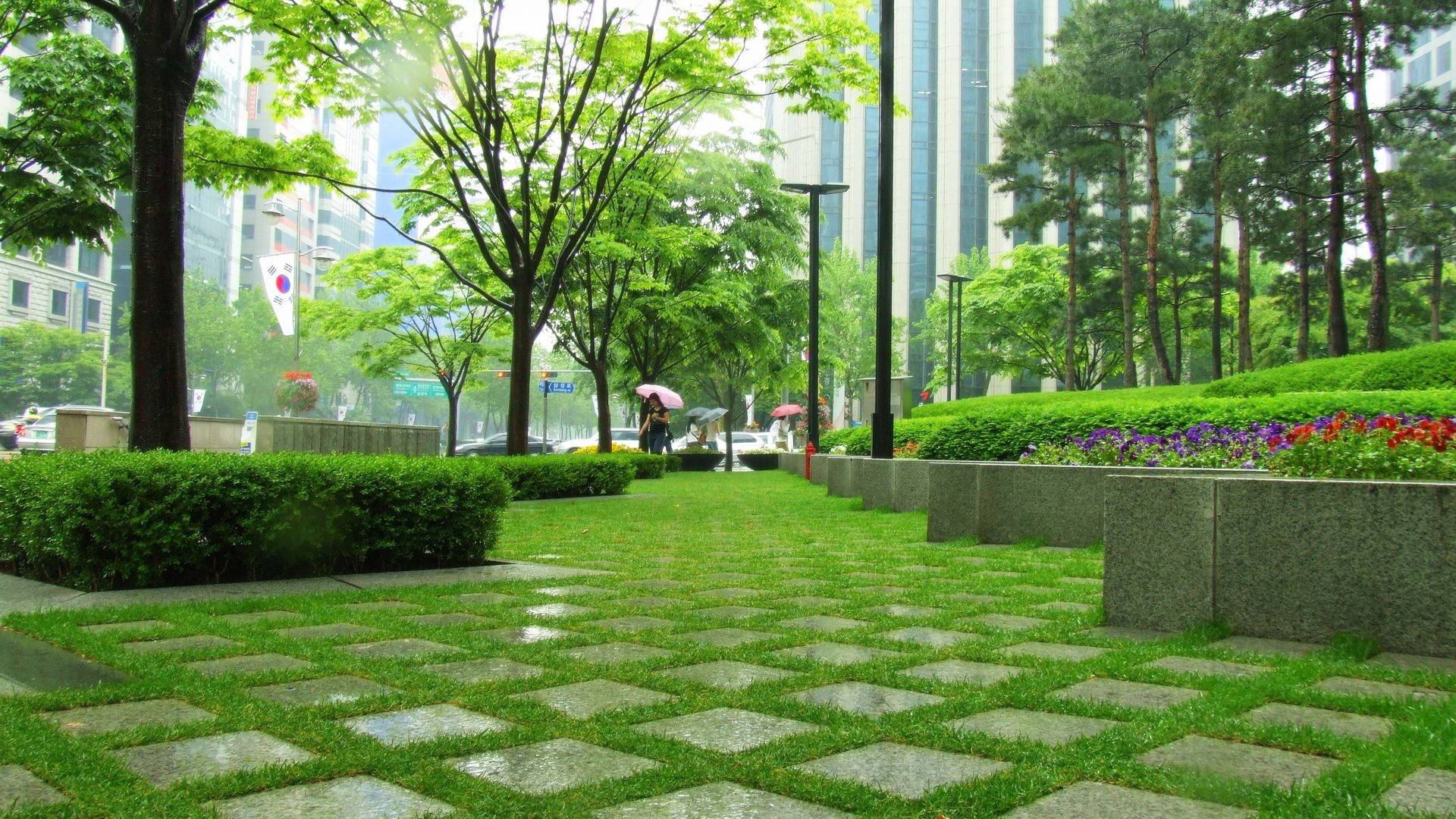 德黑蘭綠色步道 壁紙 — 一個被雨水淋濕的城市公園，幾何形狀的石徑蜿蜒穿過茂密的綠草和樹木，遠處有人撐著雨傘。
