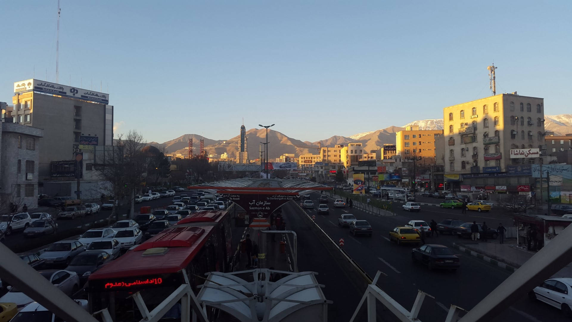 德黑蘭繁忙道路 壁紙 — 日落時分繁華的城市街道，背景是白雪覆蓋的山脈，交通在晴朗的藍天下流動。