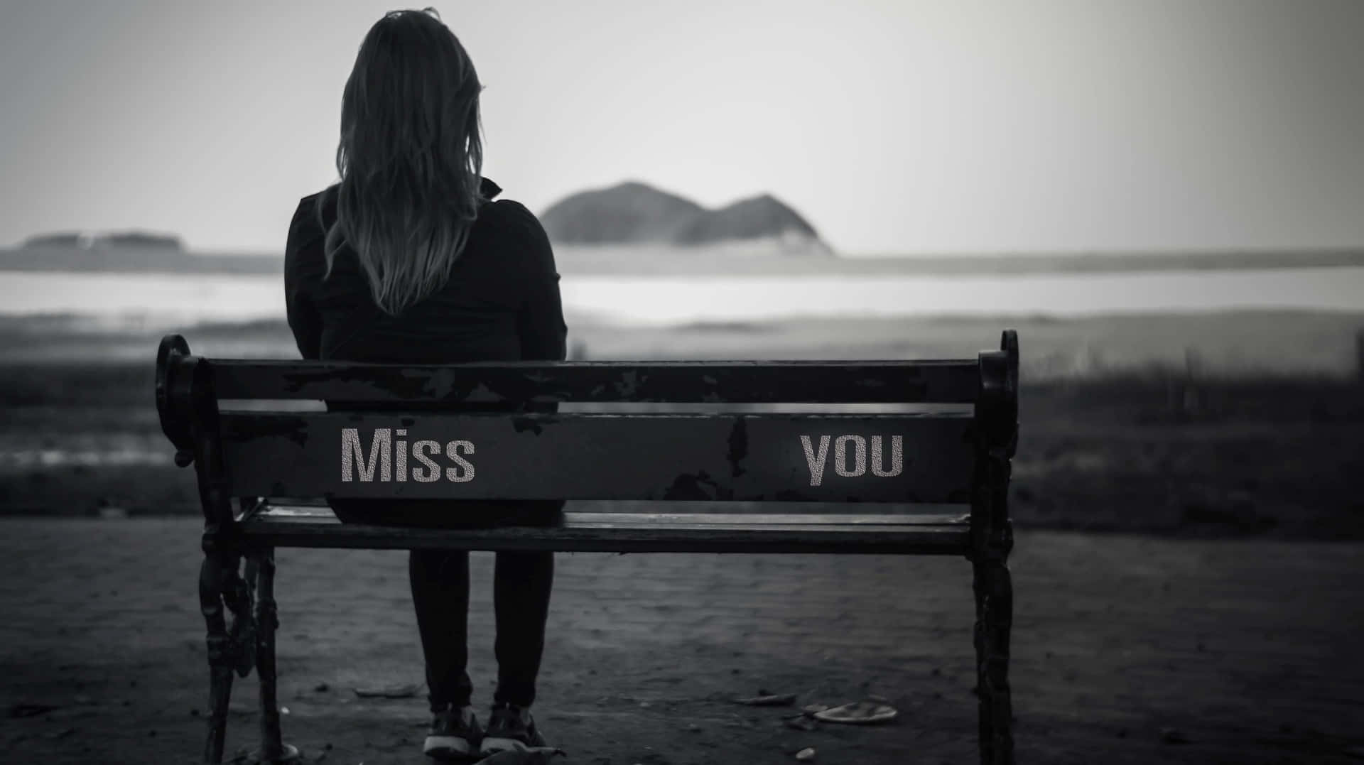 人坐在寫有 'Miss you' 的長椅上，面向水面。