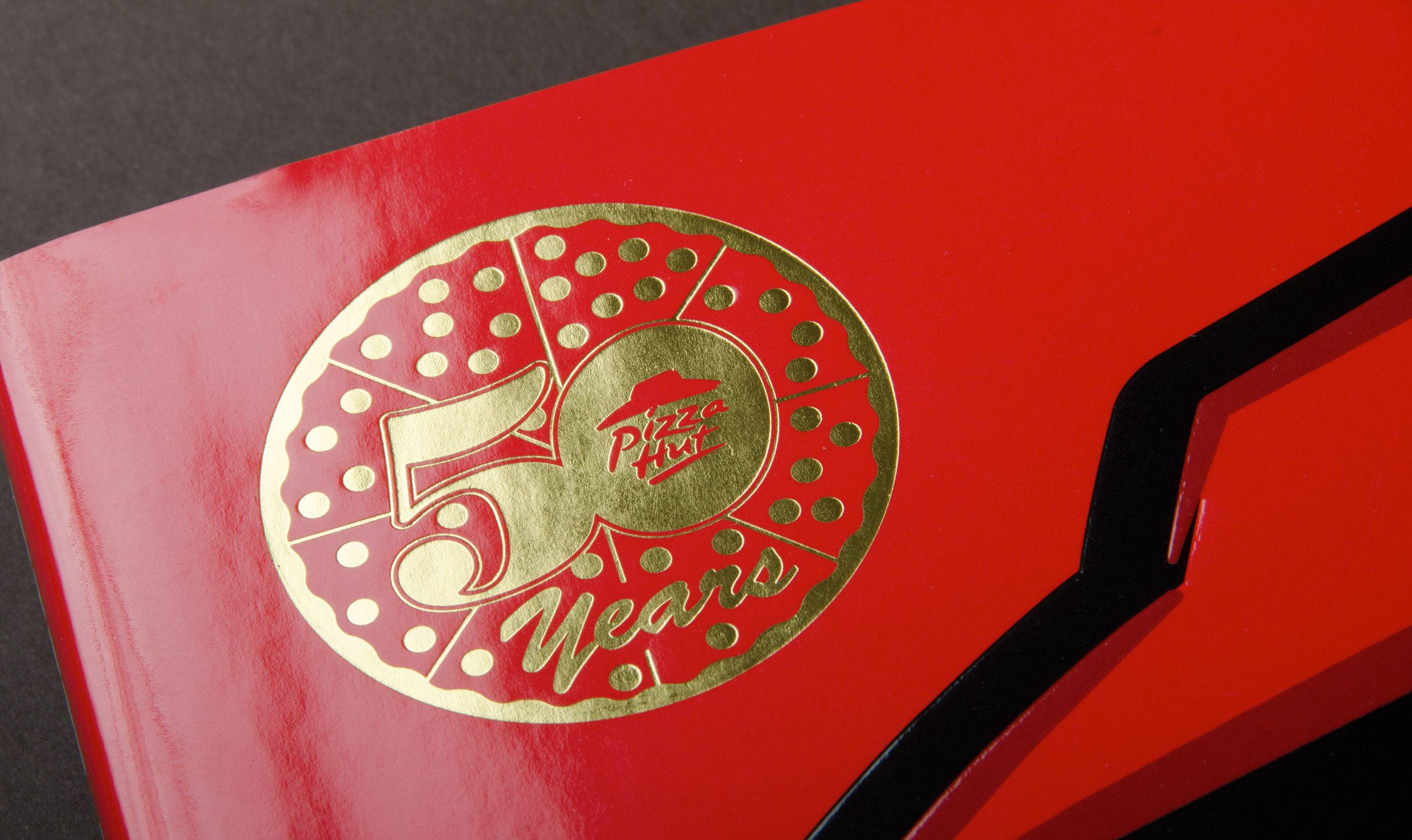 必勝客50週年故事書 壁紙 — 特寫一個亮面紅色盒子，上面有金色箔紙Pizza Hut 50 Years標誌，附有薄餅片設計。