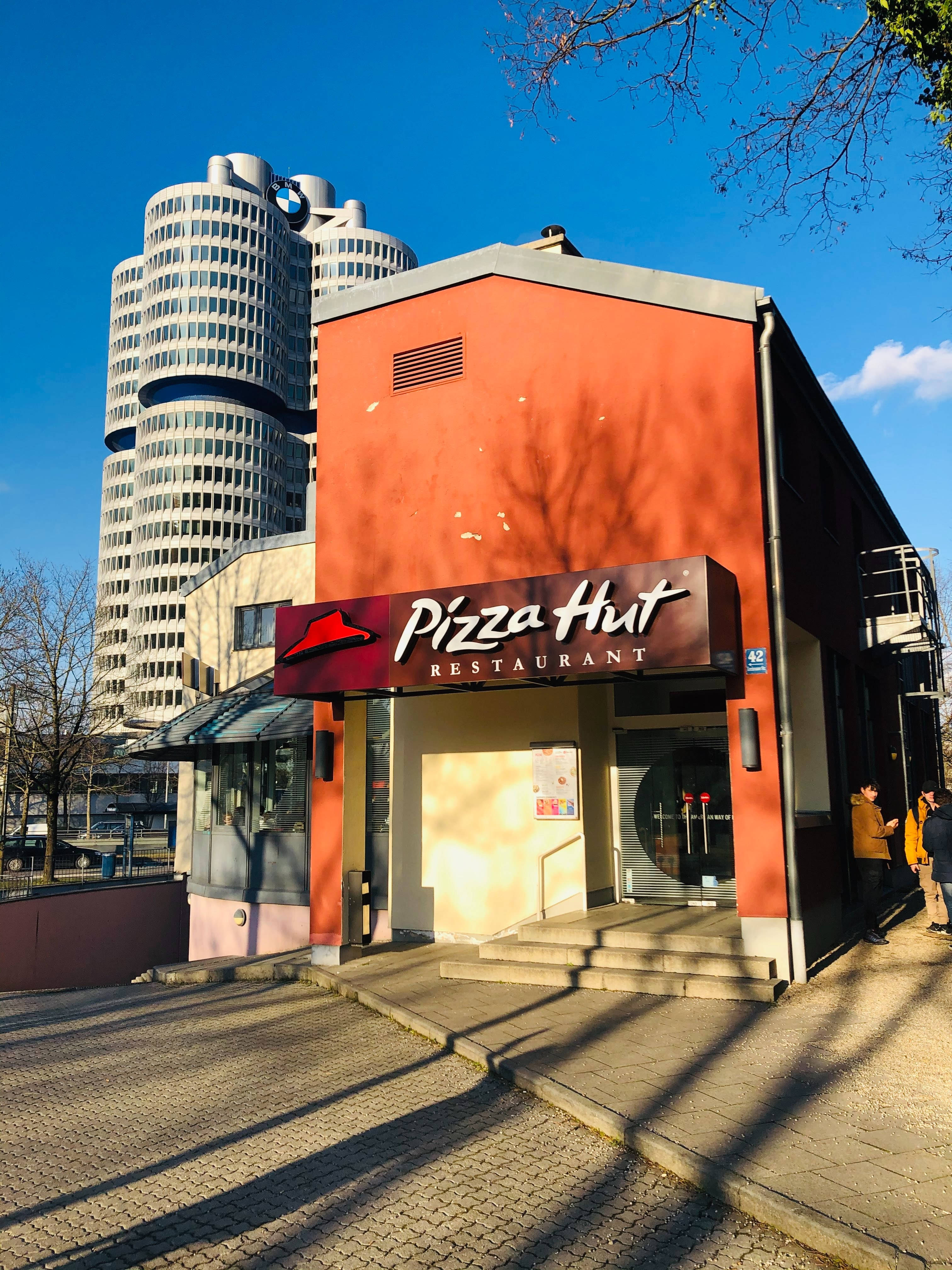 必勝客餐廳外觀 壁紙 — 一間亮橙色的必勝客餐廳矗立在標誌性的寶馬總部大樓前，天空湛藍清澈。