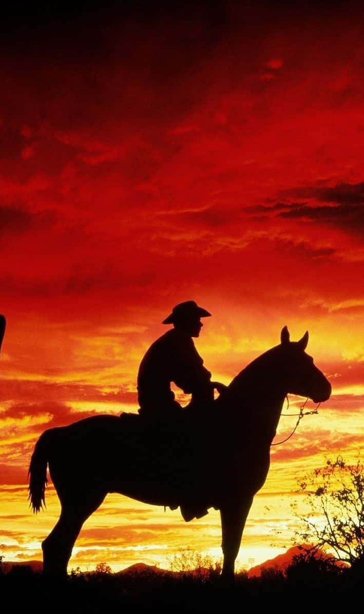 快來看看這款超酷的 Cowboy iPhone 壁紙！ 壁紙 — 一個孤獨的牛仔騎在馬背上，在火紅和橙色的日落天空襯托下形成剪影。