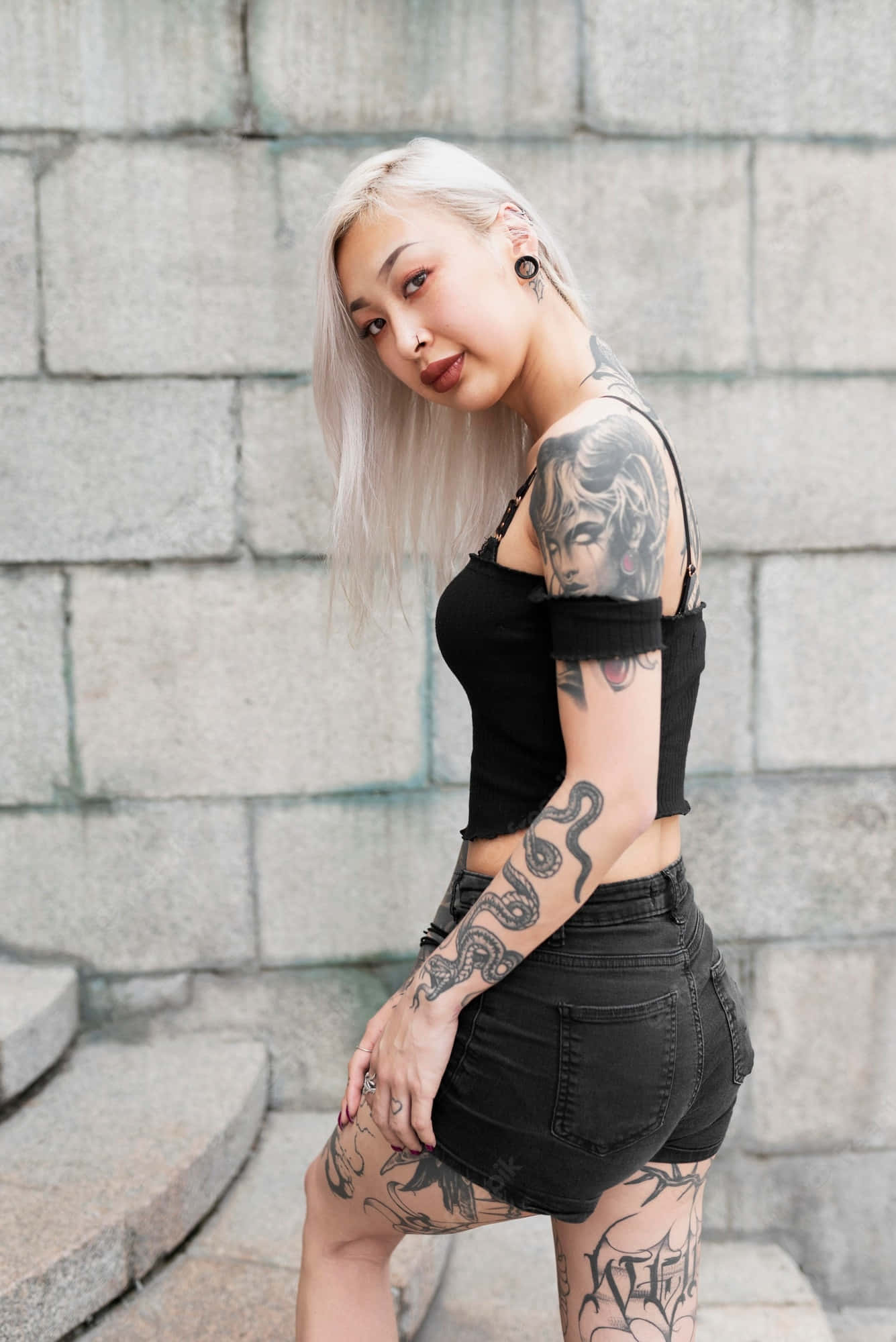 性感紋身女孩 壁紙 — 一位紋身女子，一頭鉑金色頭髮，身穿黑色短上衣和短褲，自信地靠在灰色石牆上擺姿勢。
