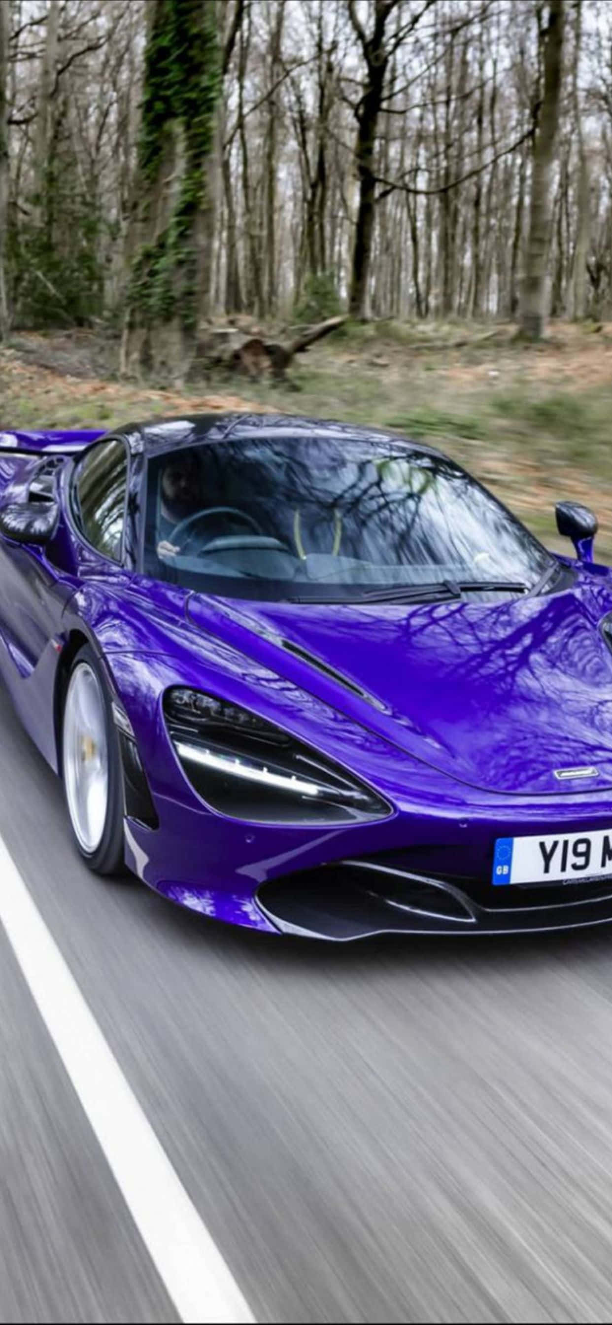 性能與奢華的完美結合——iPhone Xs Max McLaren 720S 壁紙 — 一輛充滿活力的紫色McLaren跑車沿著森林道路飛馳，背景有動態模糊和光禿的樹木。