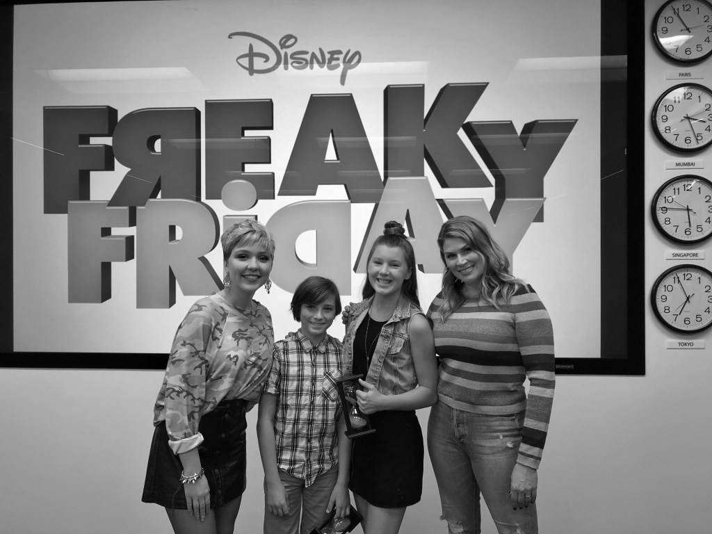 《怪誕星期五》迪士尼演員黑白照 壁紙 — 四個微笑嘅人喺一個大型嘅『Freaky Friday』迪士尼標誌前面擺甫士，牆上有全球時鐘。