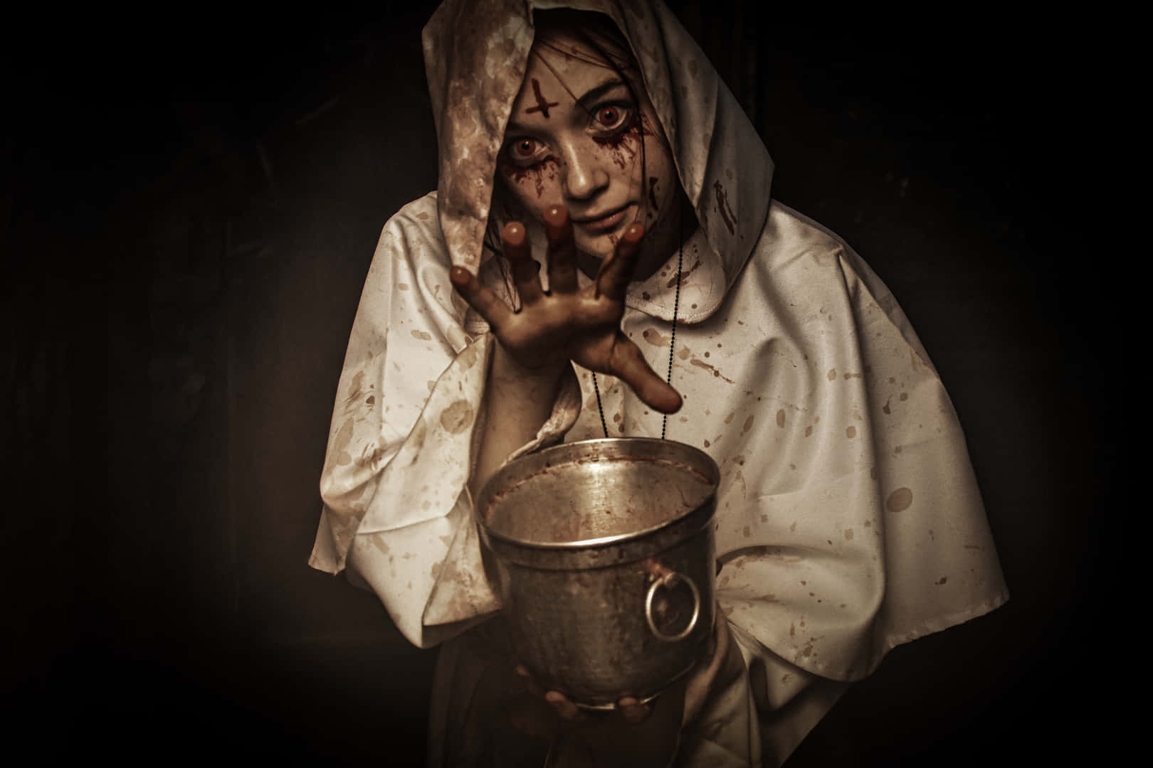 恐怖女巫畫面 壁紙 — 一個血淋淋的修女，穿著沾污的白色修女服，拿著一個金屬水桶，在黑暗、不祥的環境中舉起一隻血淋淋的手。
