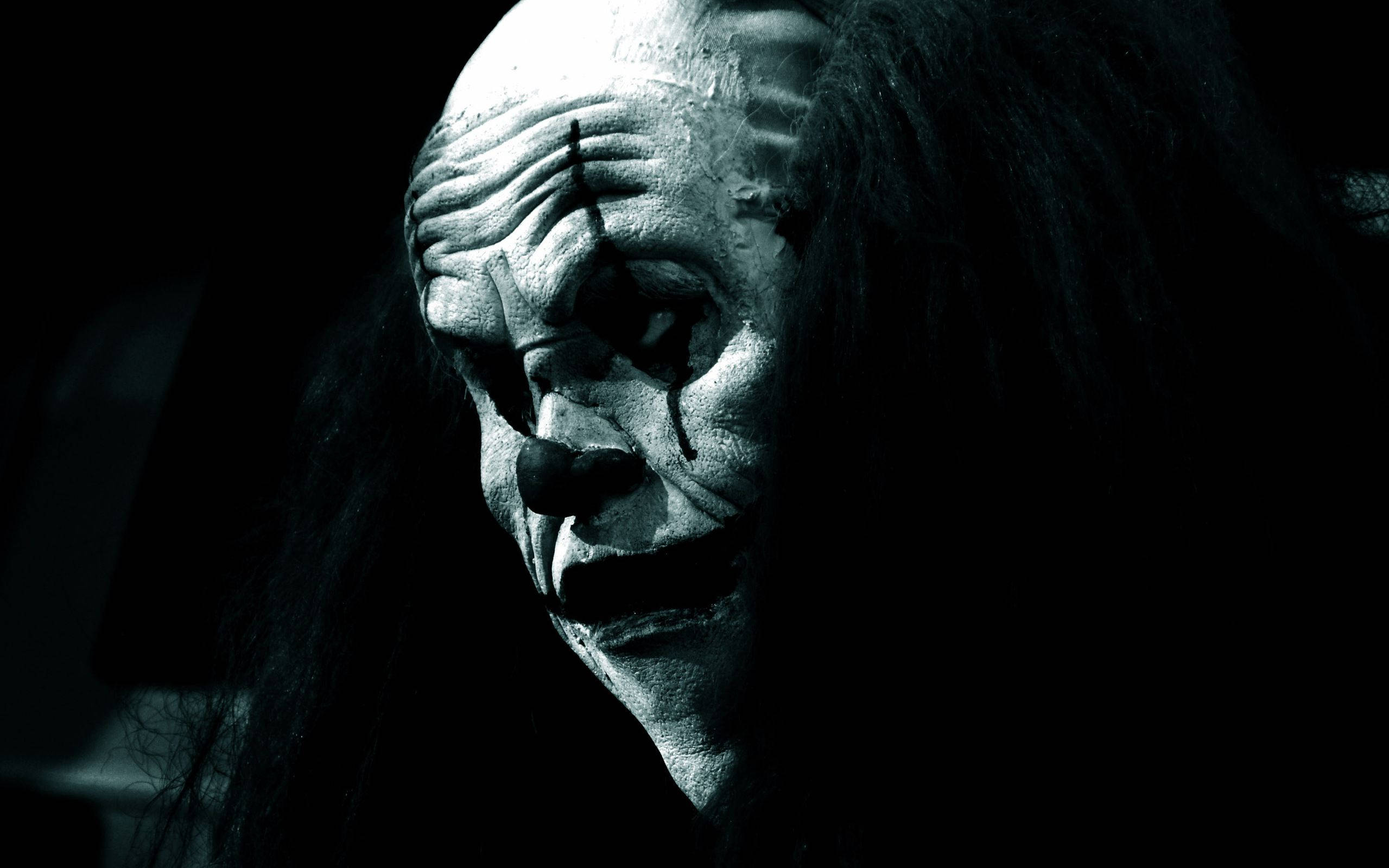 恐怖萬聖節惡魔小丑 壁紙 — 一個令人毛骨悚然的小丑特寫，臉上帶著裂開的白色化妝和黑色淚水，在昏暗的環境中流淌。