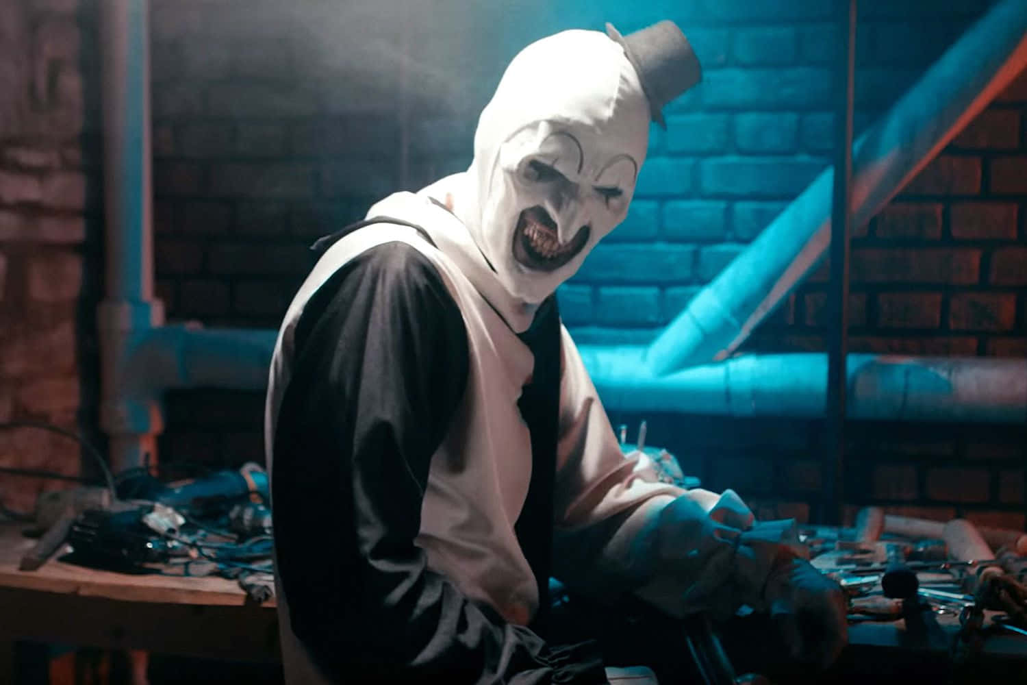 恐怖電影《Terrifier》中的小丑 Art 圖像 壁紙 — 一個戴著白色面具和高帽的威脅性小丑站在昏暗的磚房裡，周圍是工具和藍色燈光。