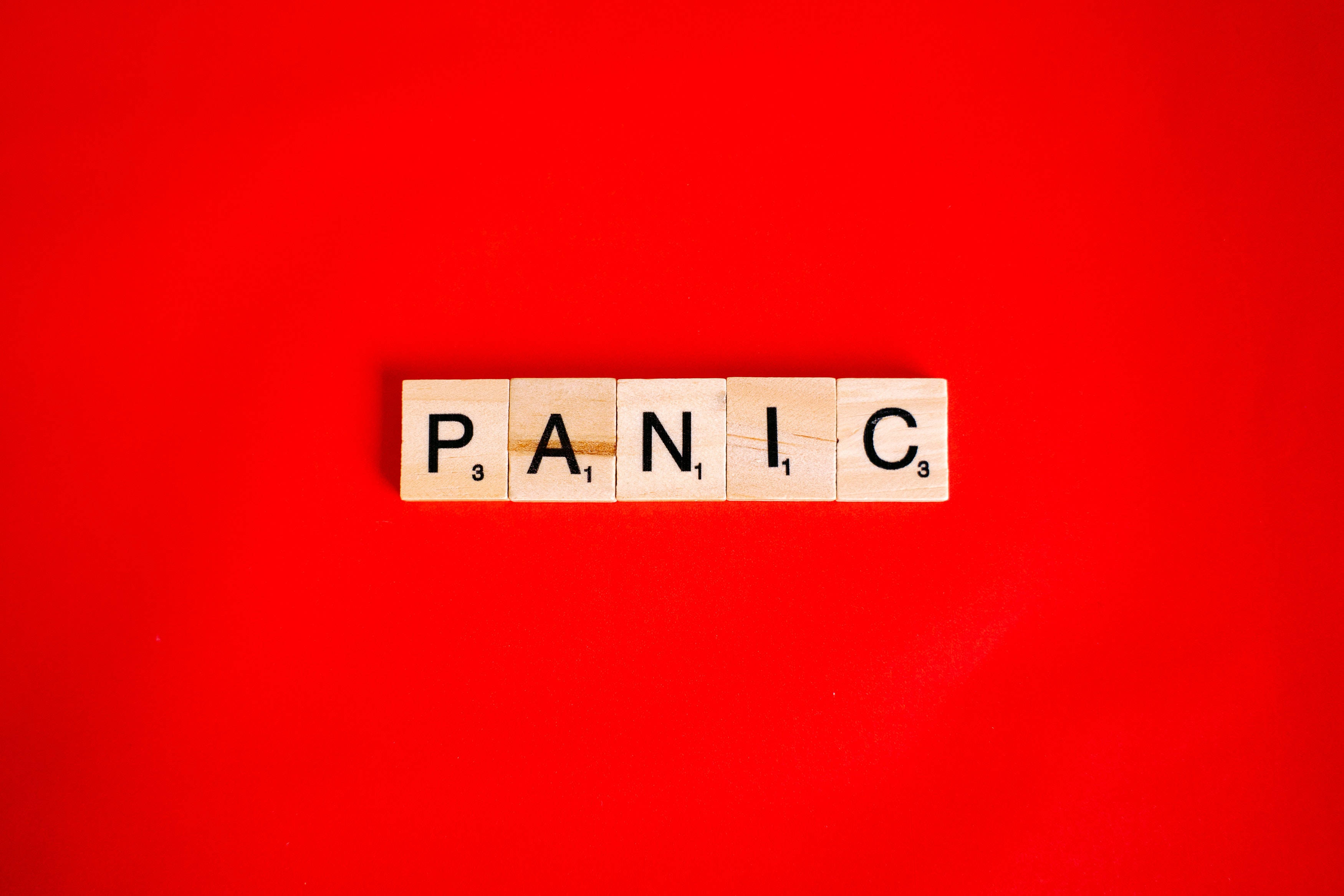 「恐慌」拼字遊戲方塊 壁紙 — 單詞「PANIC」用木製Scrabble字母塊拼出，居中放在鮮紅色背景上。