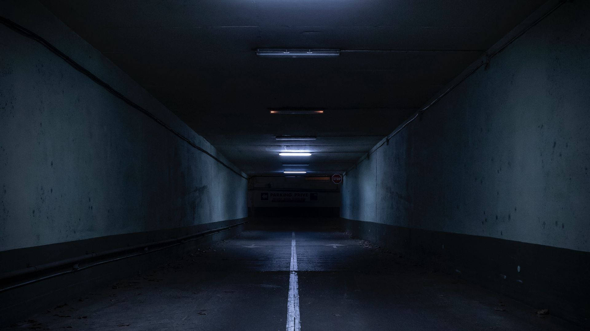 恐懼的黑暗地下隧道 壁紙 — 一條長而昏暗的地下停車場隧道，中央有一條白線，頭頂上有閃爍的螢光燈。