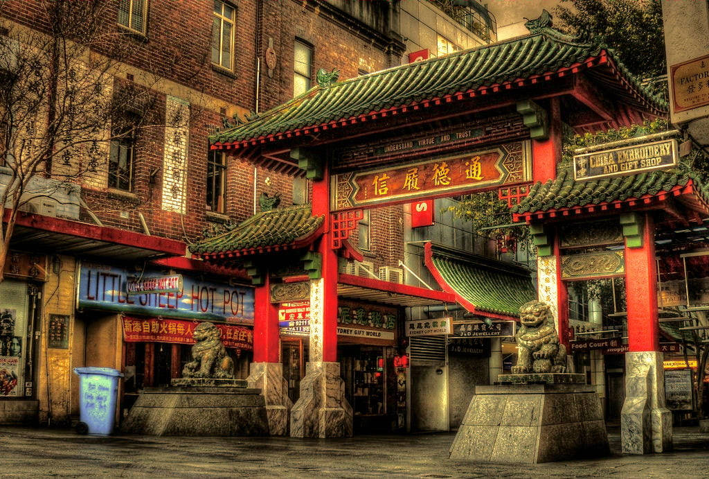 悉尼唐人街迪克遜街 壁紙 — 一座充滿活力的紅綠中國牌坊，配以石獅，矗立於繁華的唐人街街道入口，兩旁是磚砌建築和發光的招牌。