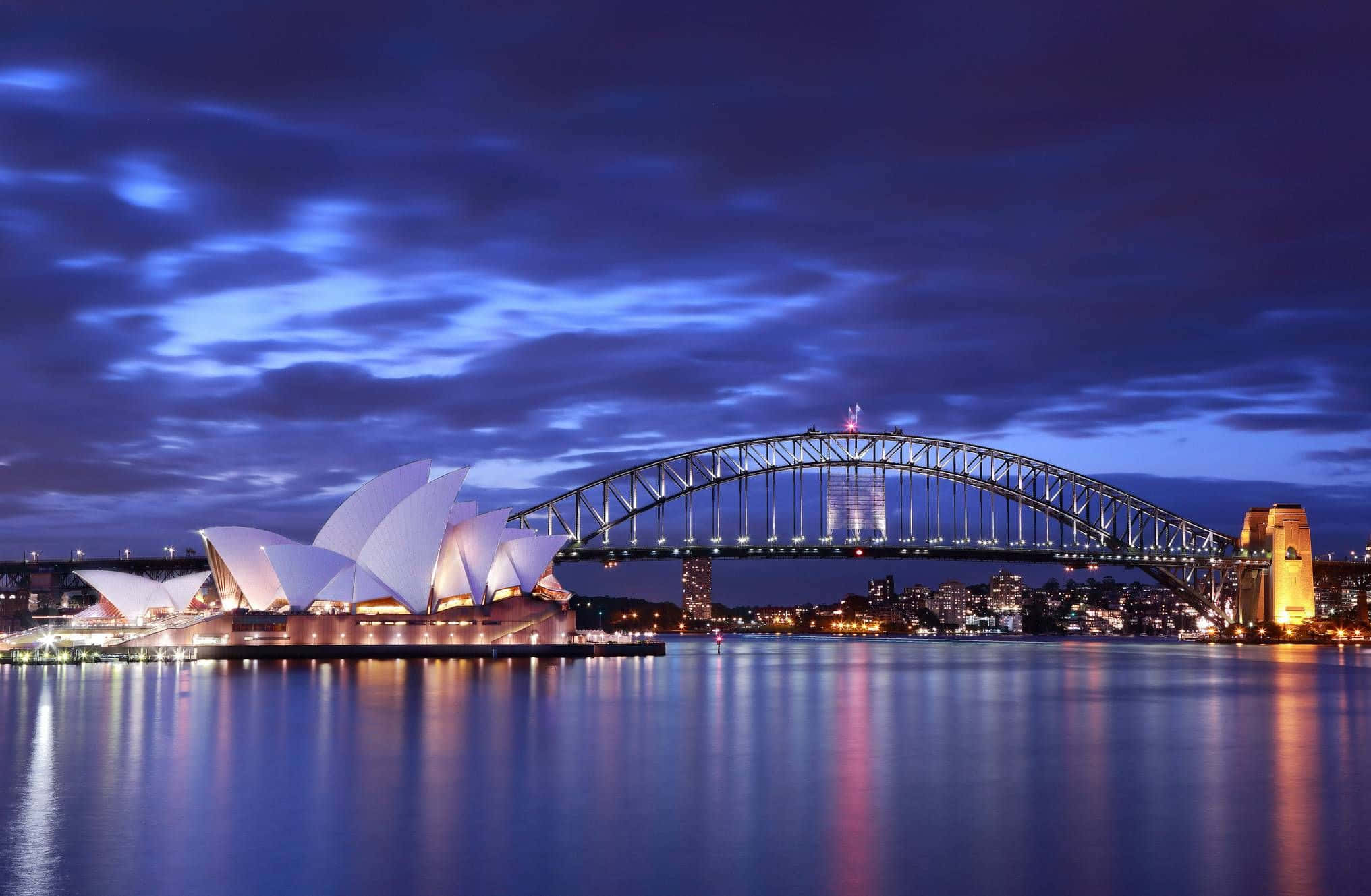 悉尼歌劇院日落美景 壁紙 — 悉尼歌劇院和海港大橋在黃昏時亮起燈光，倒影在平靜的水面上閃爍，深藍色的天空下。