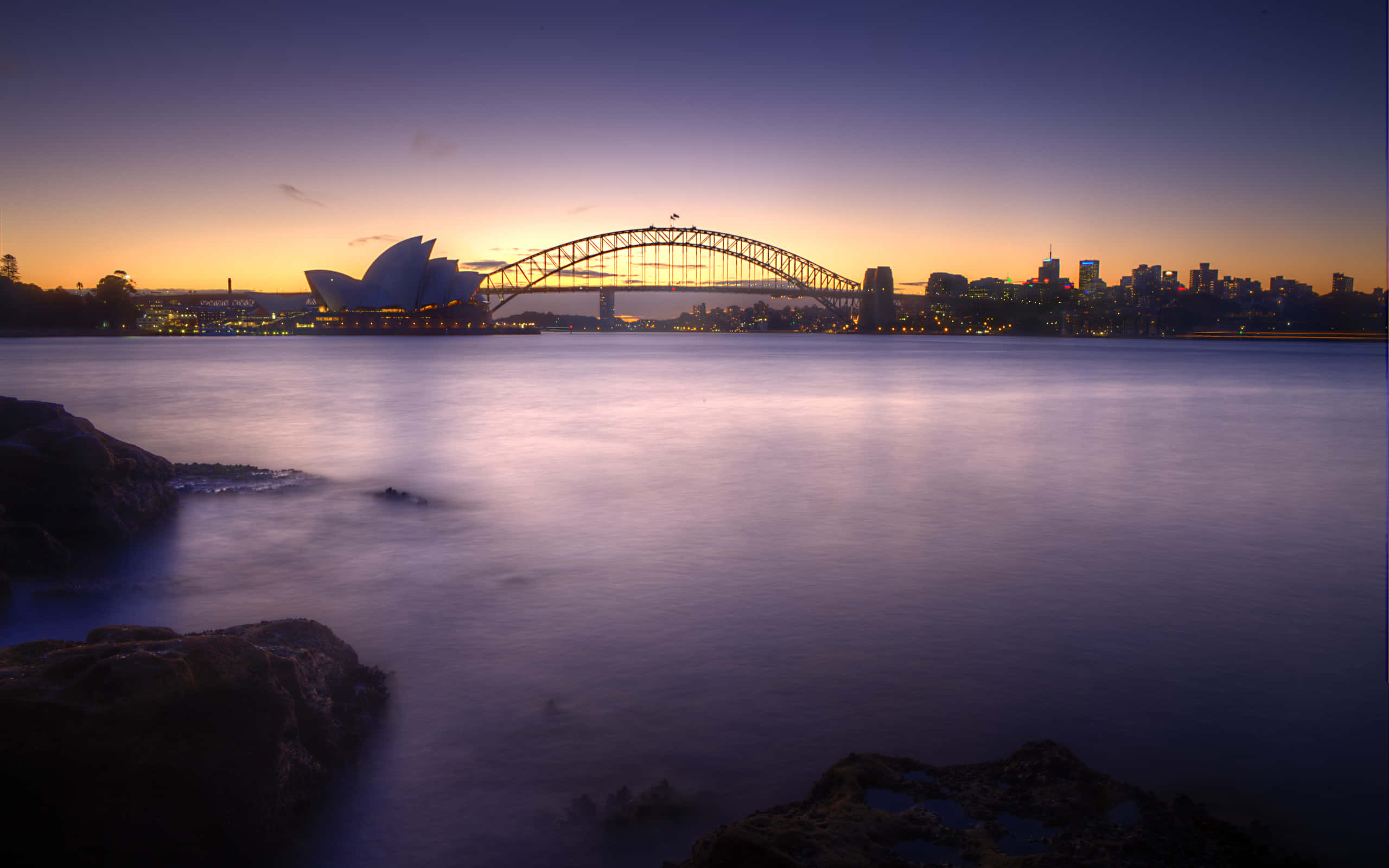 Australia 桌布 壁紙 — 悉尼港灣大橋和歌劇院在黃昏時分閃耀，平靜的水面和城市燈光映照在寧靜的海港中。