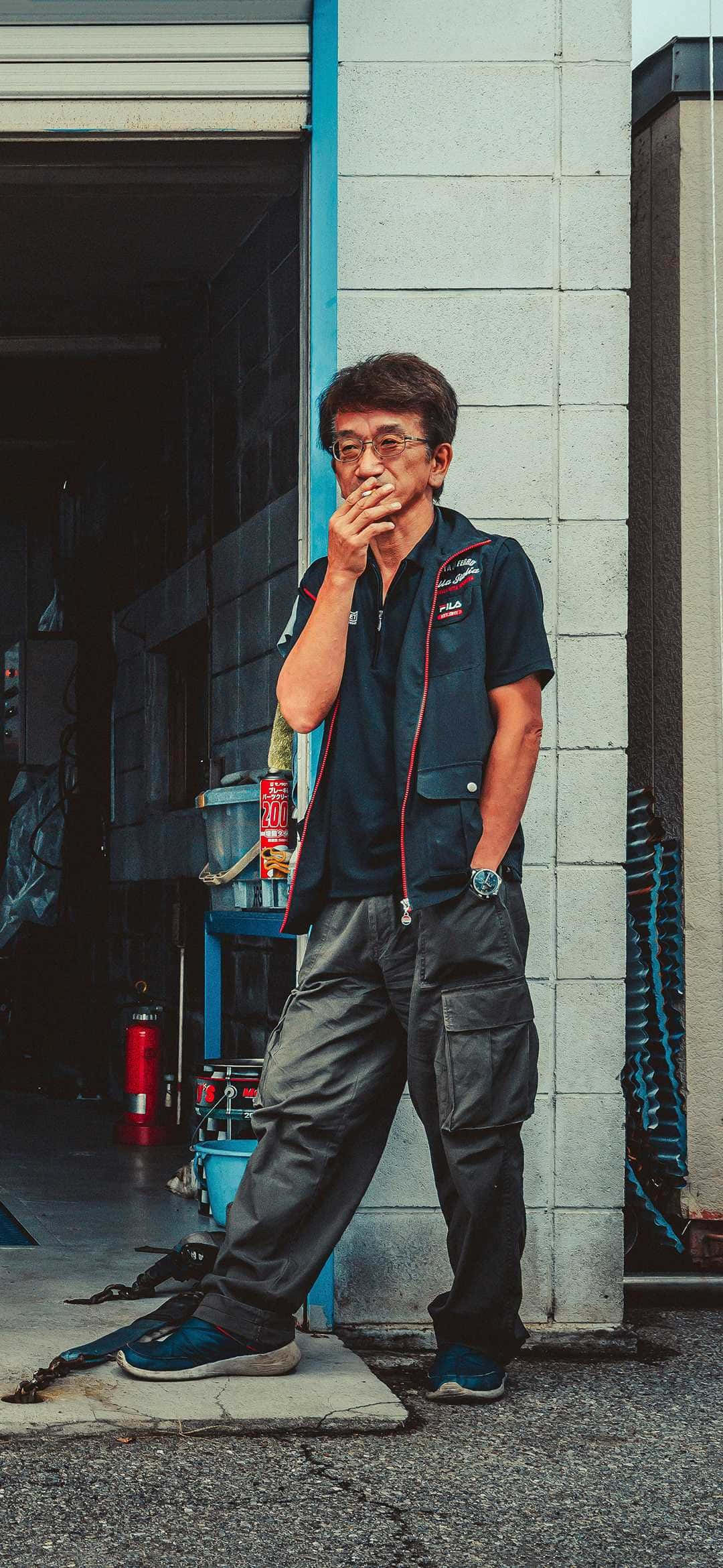 悠閒站立的煙霧 Nagata 壁紙 — 一位中年機械師穿著深色背心和工裝褲，靠在車庫牆上抽煙。