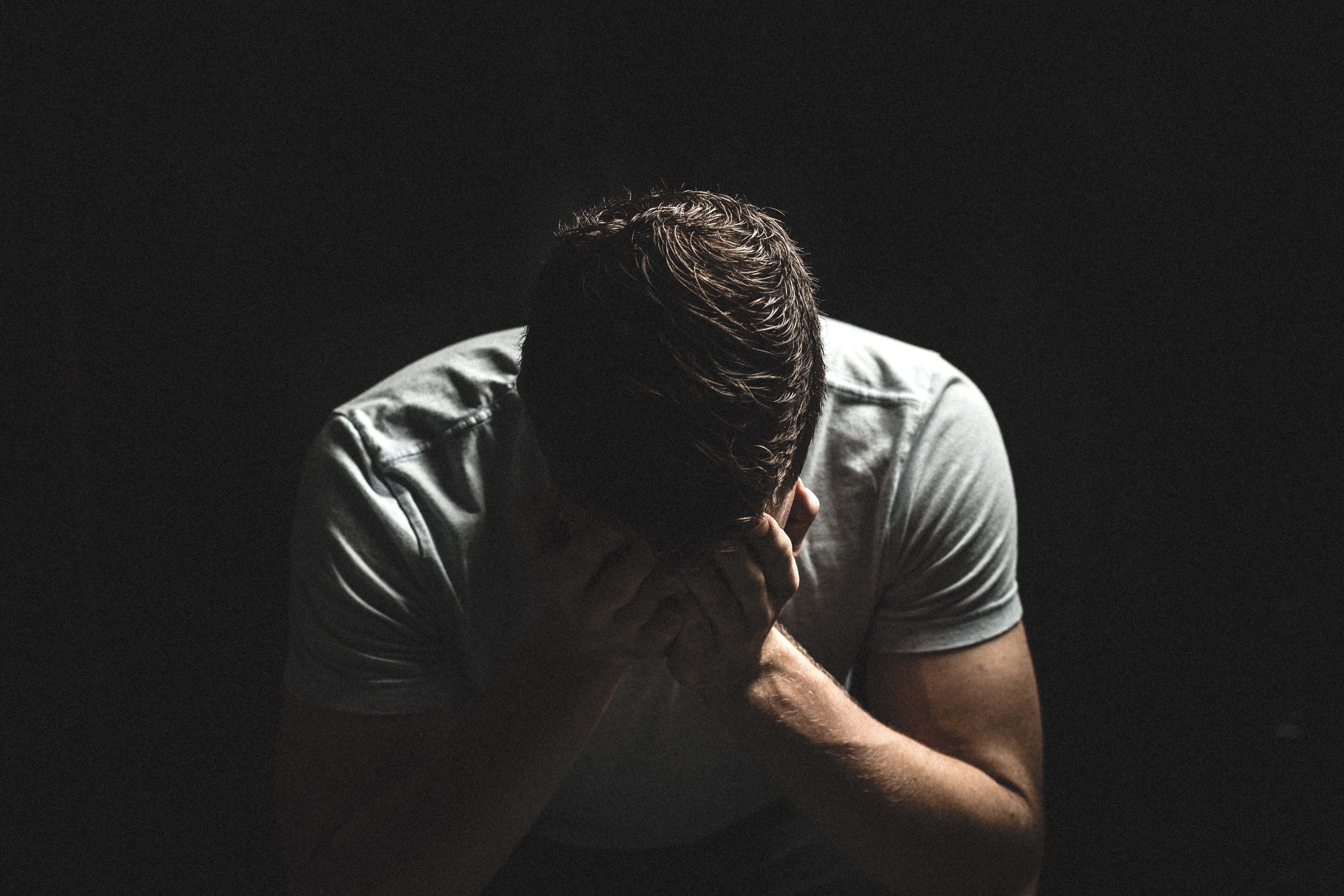 悲傷男子拭淚圖片 壁紙 — 一名身穿白色T恤的男子在純黑背景下彎腰低頭，雙手抱頭。