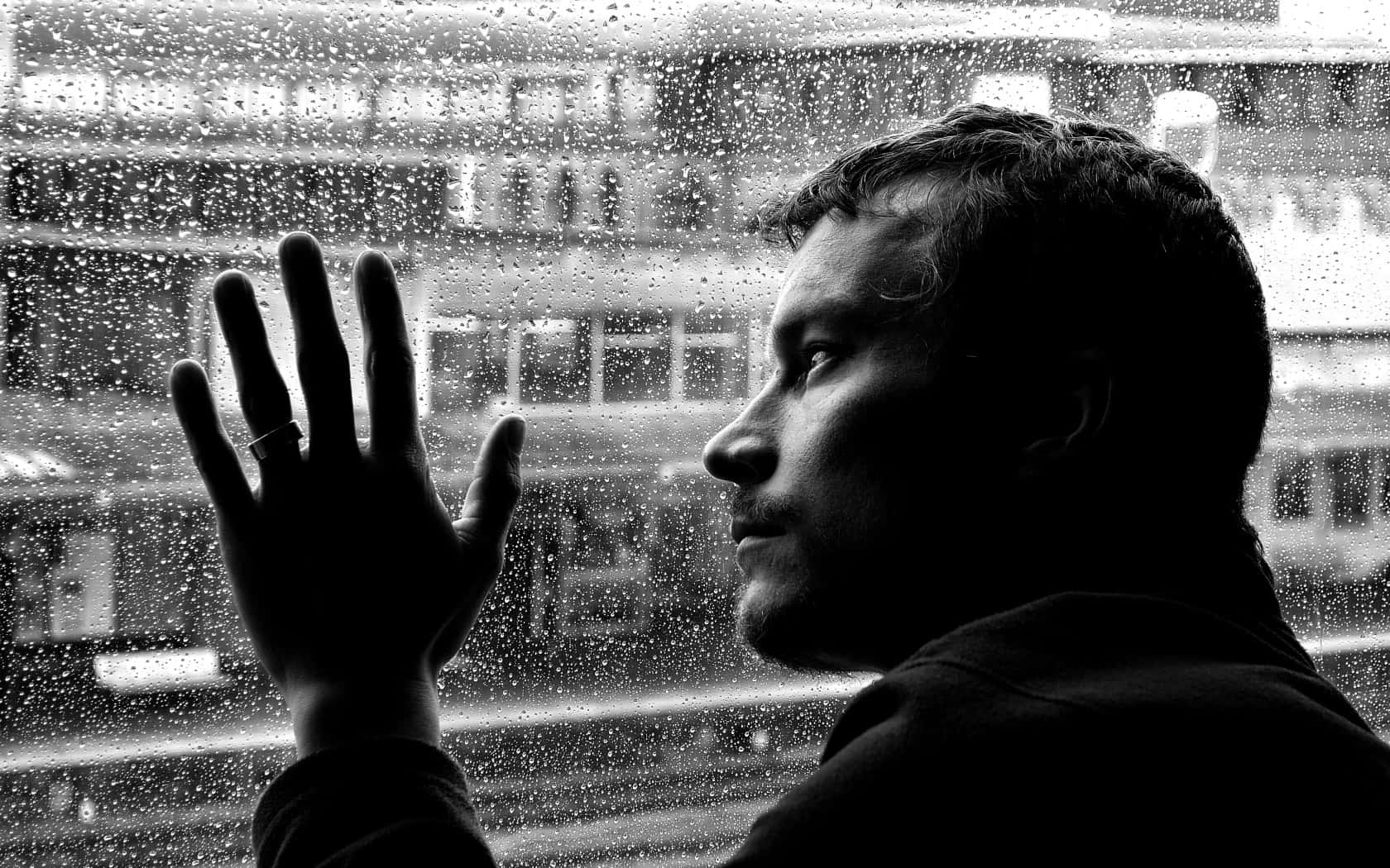 悲傷男子窺視窗外 壁紙 — 一個剪影中的男人透過佈滿雨點的窗戶凝視著單色調模糊的城市景觀。