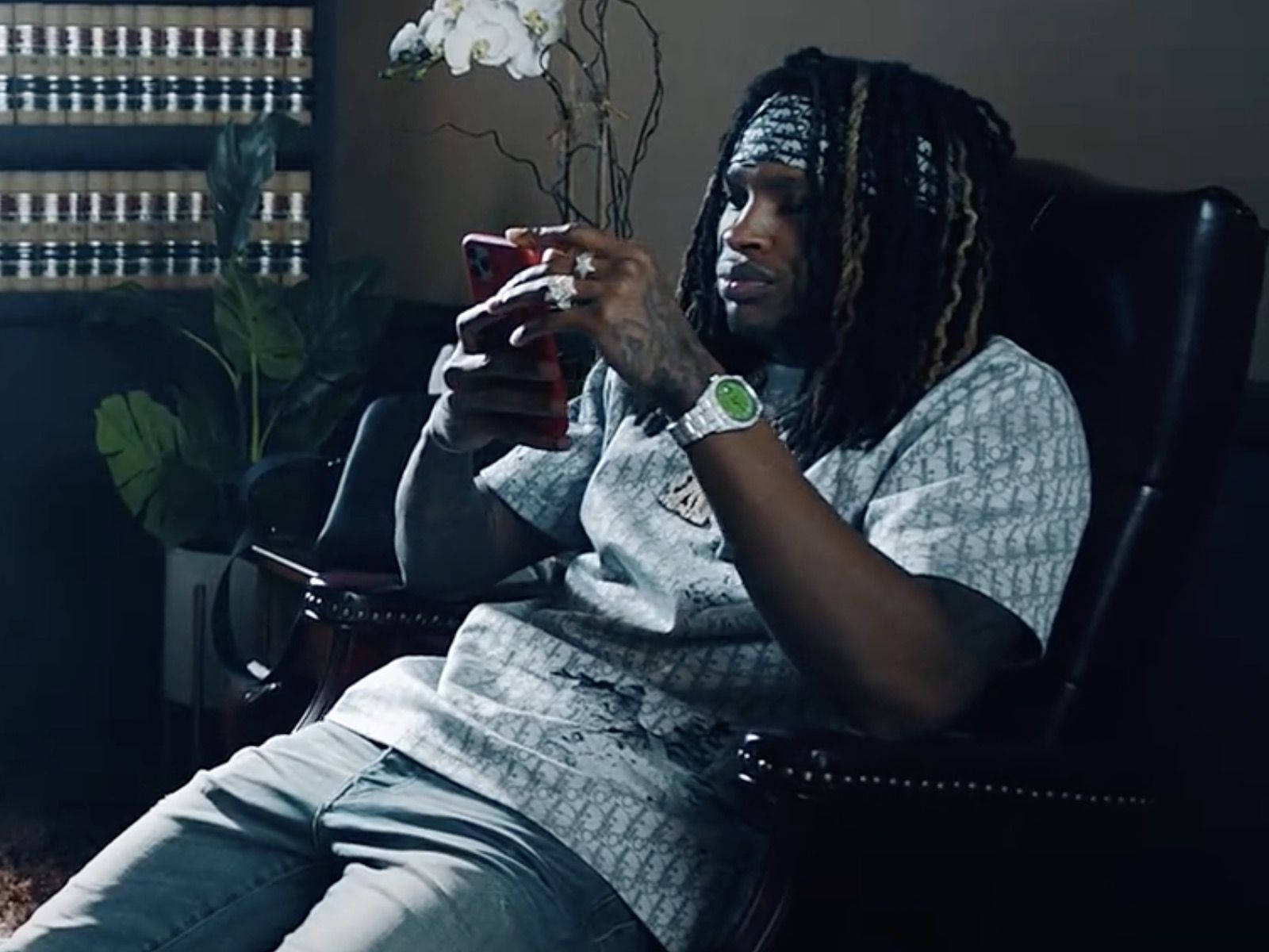 悼念 King Von 坐著沉思 壁紙 — 一名留著辮子的男人坐在皮椅上，在昏暗的房間裡拿著紅色電話，周圍有植物和書架。