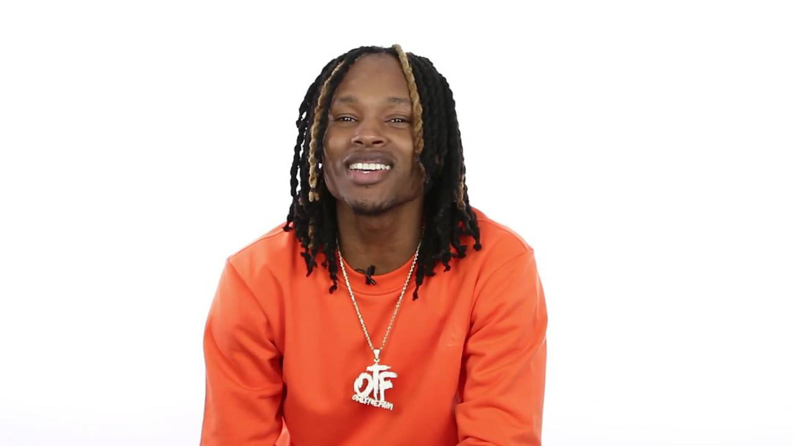 留著辮子的男人穿著橙色運動衫和項鍊對著鏡頭微笑。
