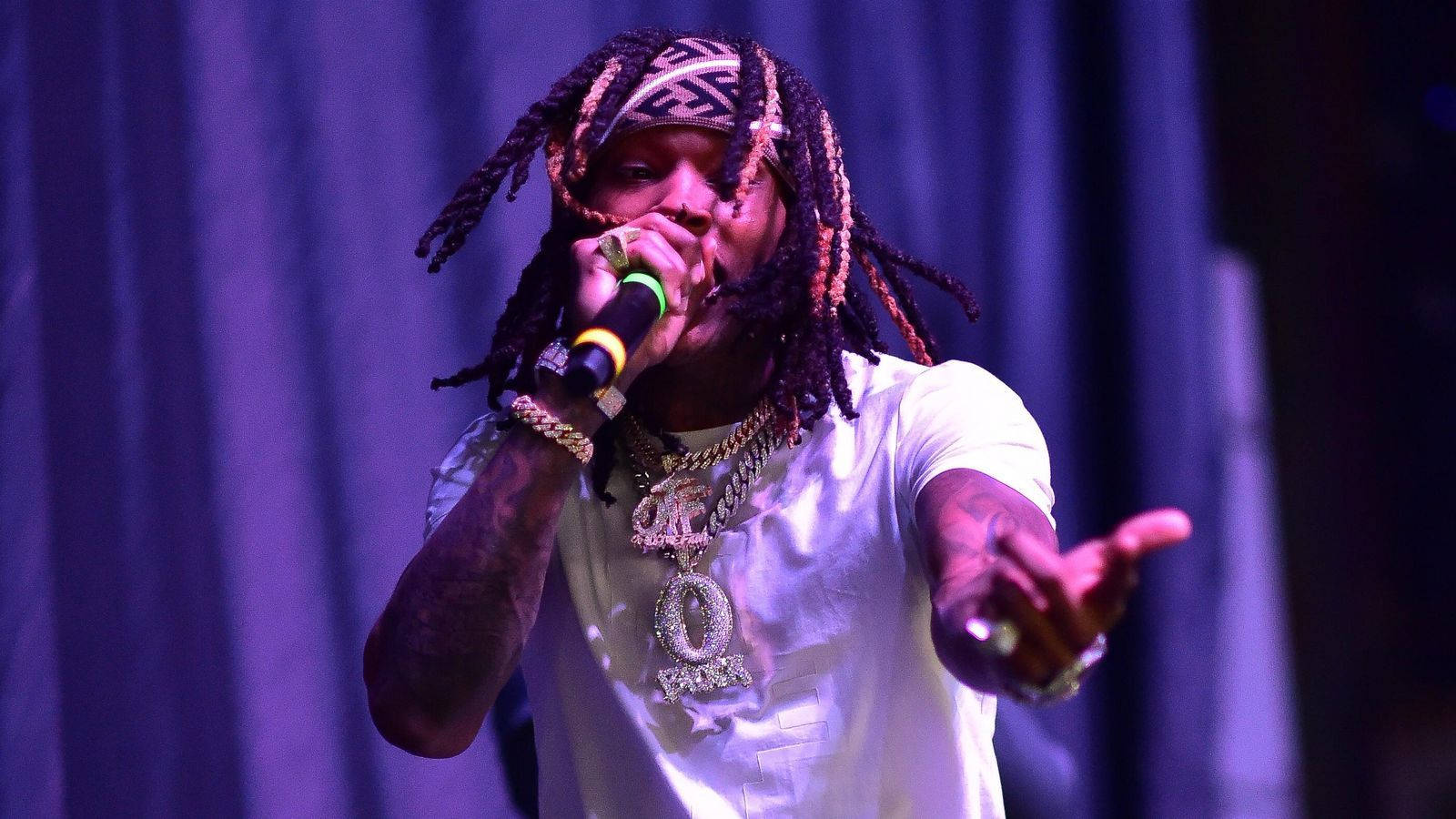 悼念 King Von 演唱持麥克風 壁紙 — 一位辮子頭髮、戴金鏈的饒舌歌手在紫色舞台燈光下熱情地對著麥克風表演。