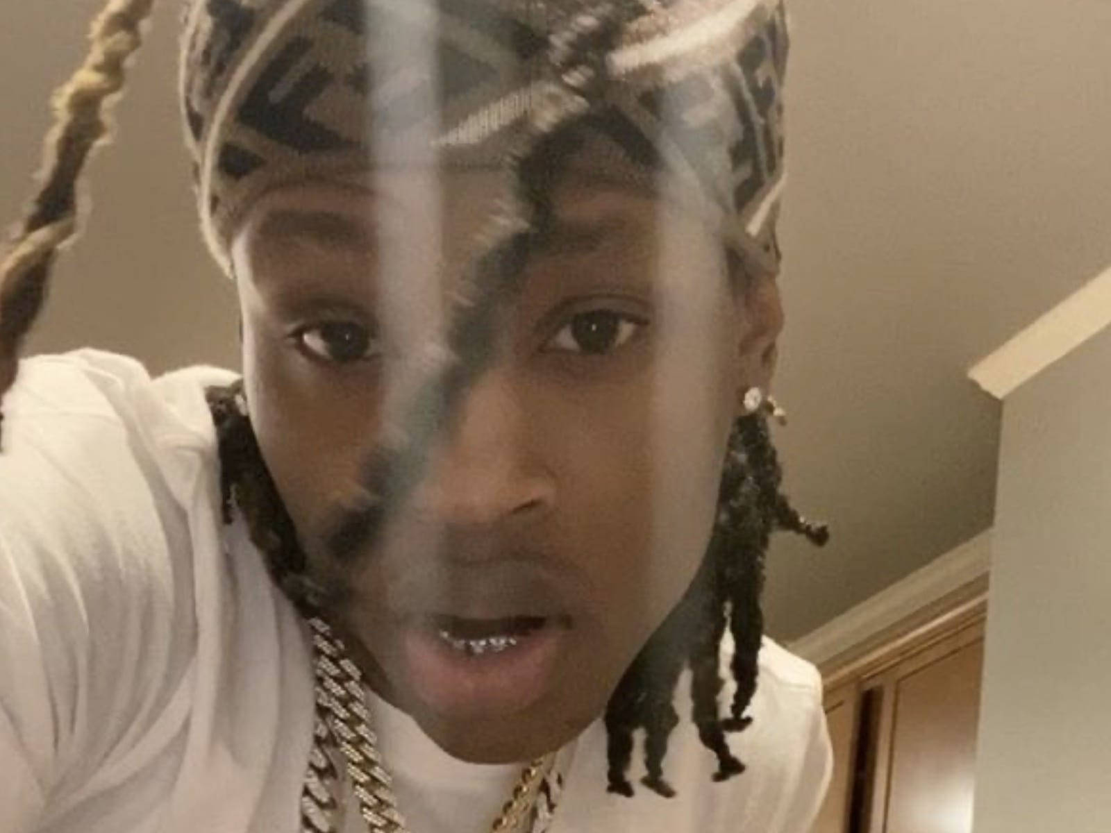 悼念 King Von 白色襯衫凝視 壁紙 — 一個辮子頭髮、戴著圖案頭巾的人，穿著白色襯衫和金色鏈條，透過反光表面觀看。