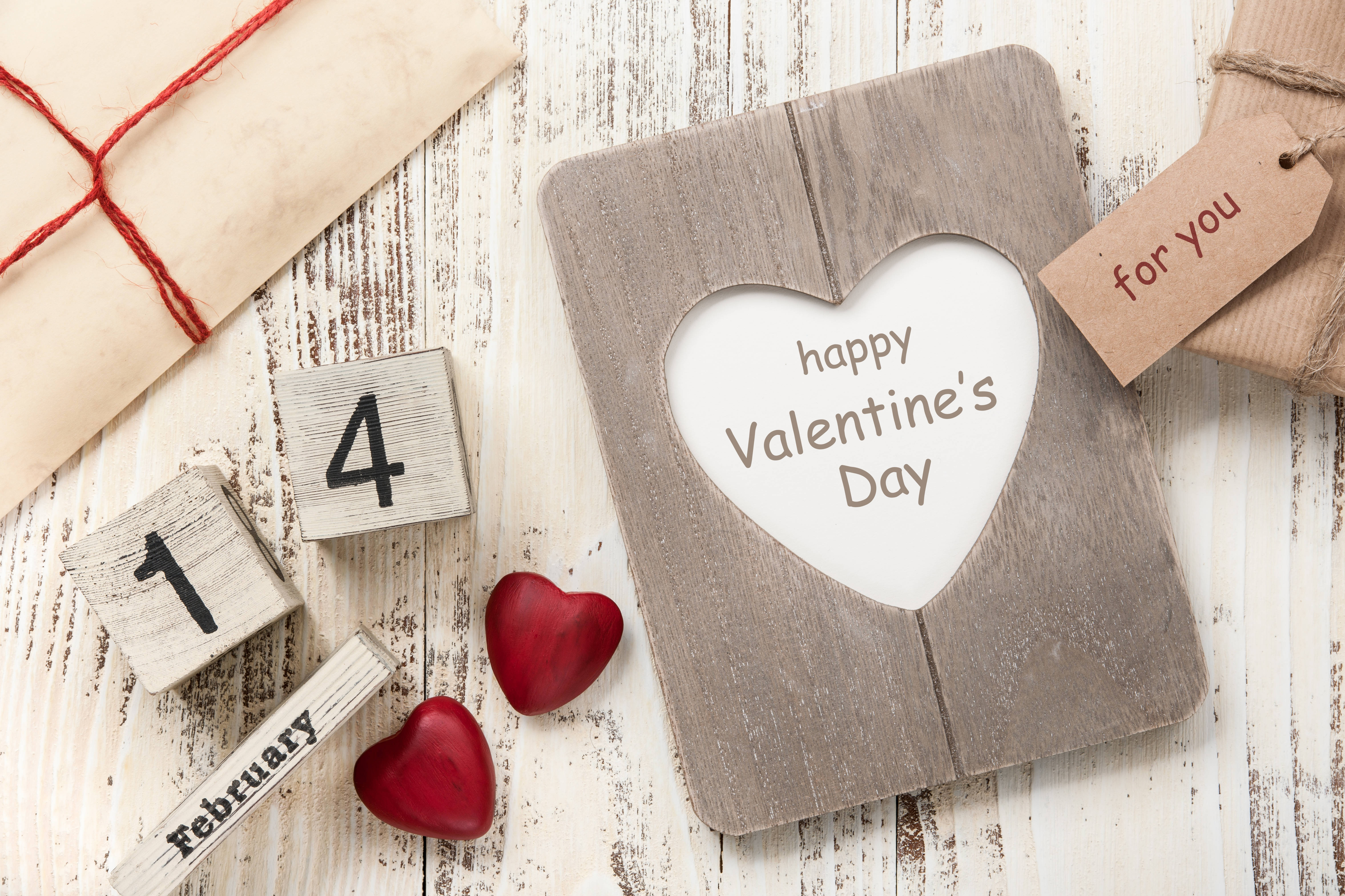 情人節二月十四 壁紙 — 一個質樸的木製心形相框，上面有「happy Valentine's Day」文字，旁邊有紅色心形、日期方塊和禮物，放在經風化的白色木質表面上。