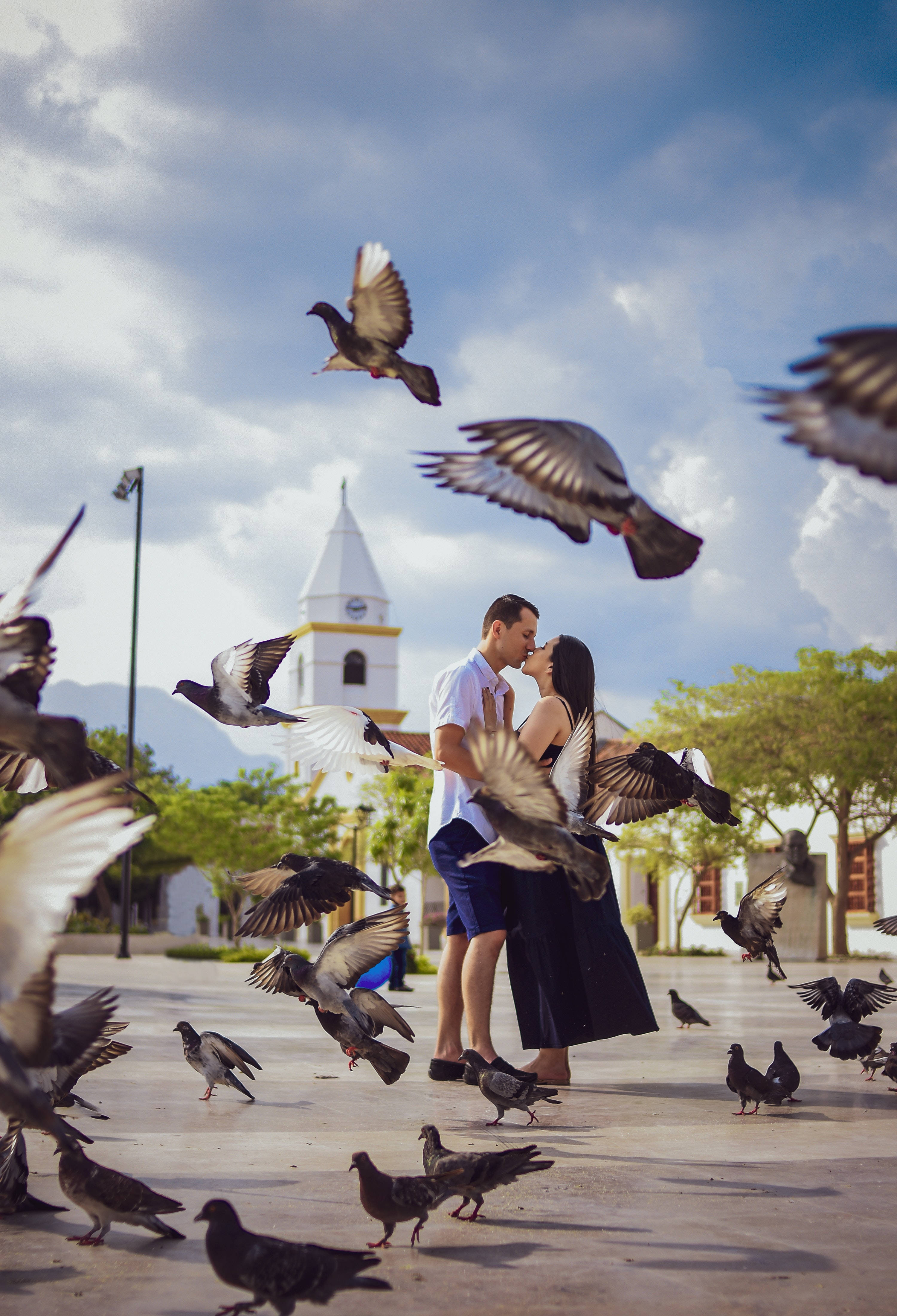 情侶公園擁吻鳥群 壁紙 — 一對情侶在陽光普照的廣場上親吻，鴿子在他們周圍飛翔並散開，附近有一座教堂塔樓。