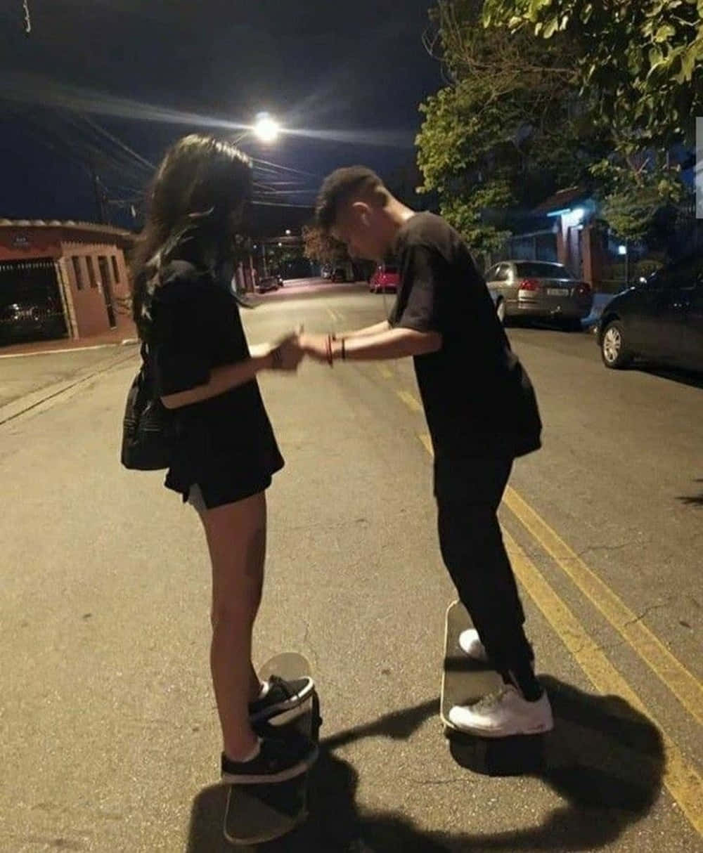 情侶夜間滑板美照 壁紙 — 兩個身穿黑色服裝的青少年在夜晚寧靜的住宅街道上，站在滑板上，在街燈下手牽手。