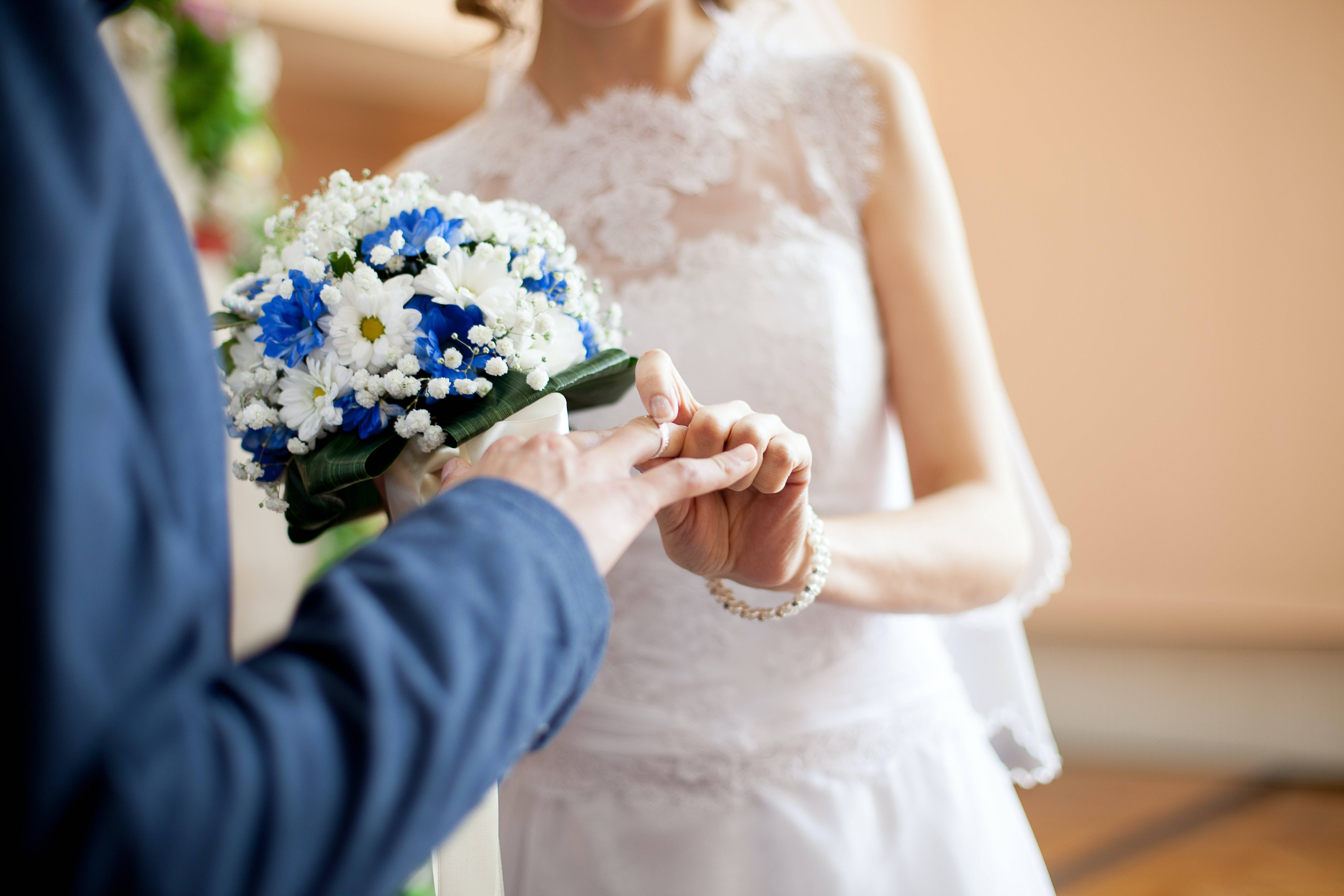 情侶手戴婚戒 壁紙 — 新郎輕輕地將結婚戒指戴在新娘的手指上，新娘手持一束鮮豔的藍白花束。