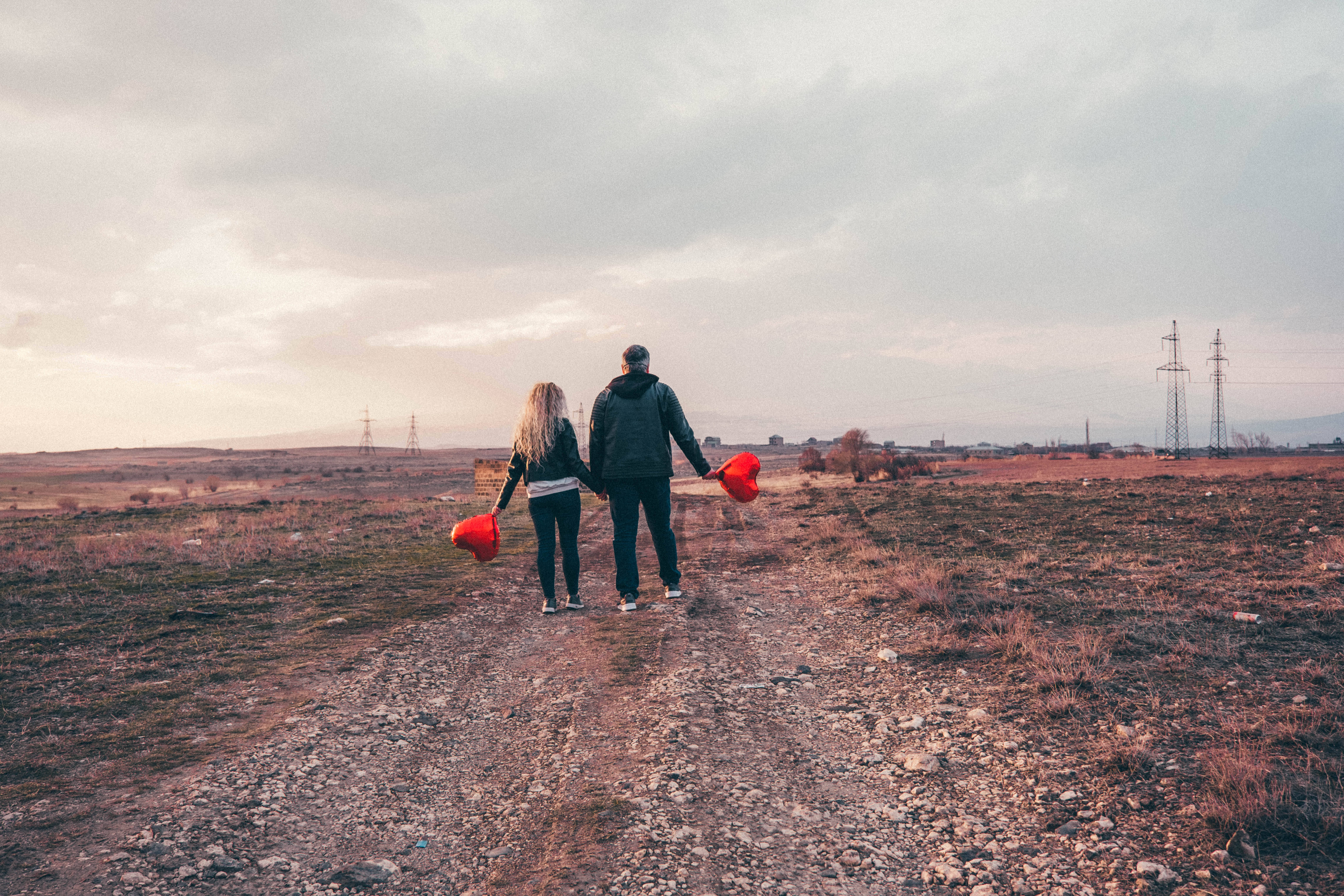 情侶攜帶可愛心形氣球 壁紙 — 一對情侶手拖手沿著碎石路穿過荒蕪的田野，在陰天中手持紅色氣球。