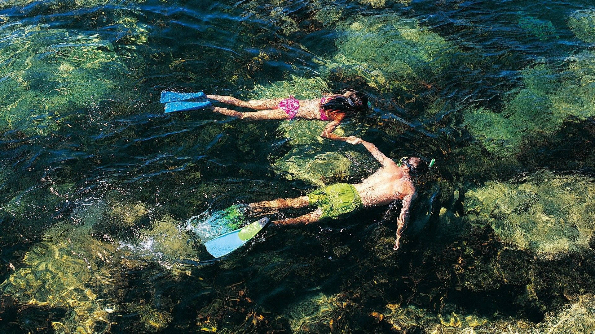 情侶潛水 壁紙 — 兩名浮潛者滑過清澈的碧綠海水，露出下方充滿活力的珊瑚和岩石。