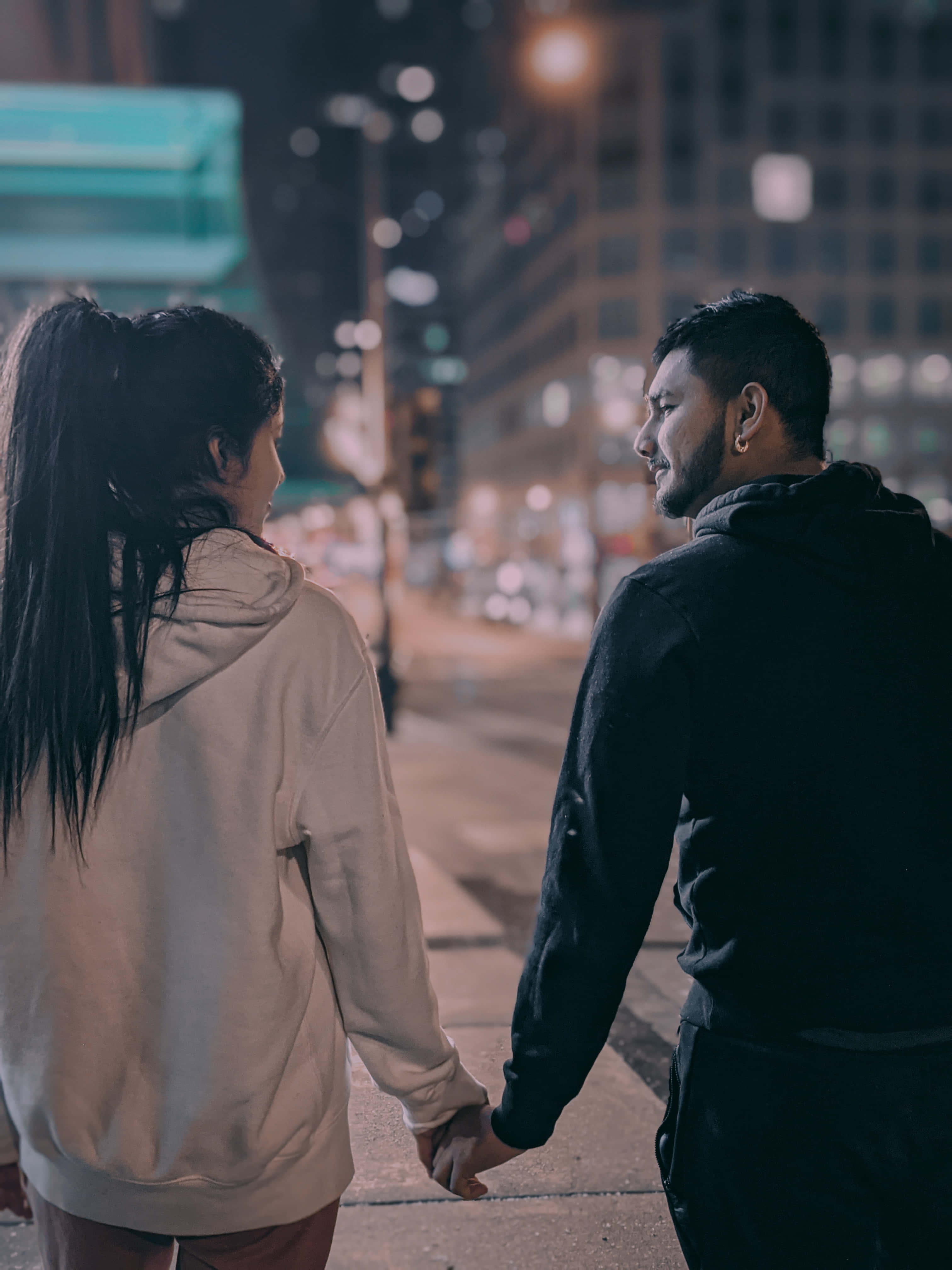 情侶牽手照 壁紙 — 一對情侶在夜晚手牽手走在城市街道上，被溫暖的路燈和身後模糊的建築燈光照亮。