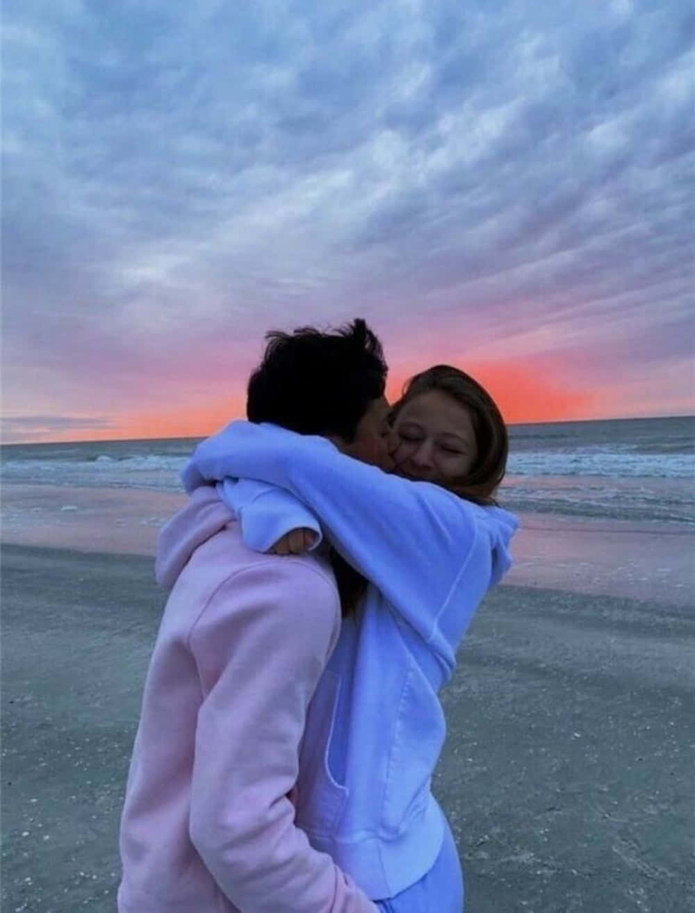 情侶穿薄荷色連帽衫夕照美圖 壁紙 — 一對情侶在沙灘上溫暖地擁抱，天空在日落時閃耀著粉紅色和橙色的色調。