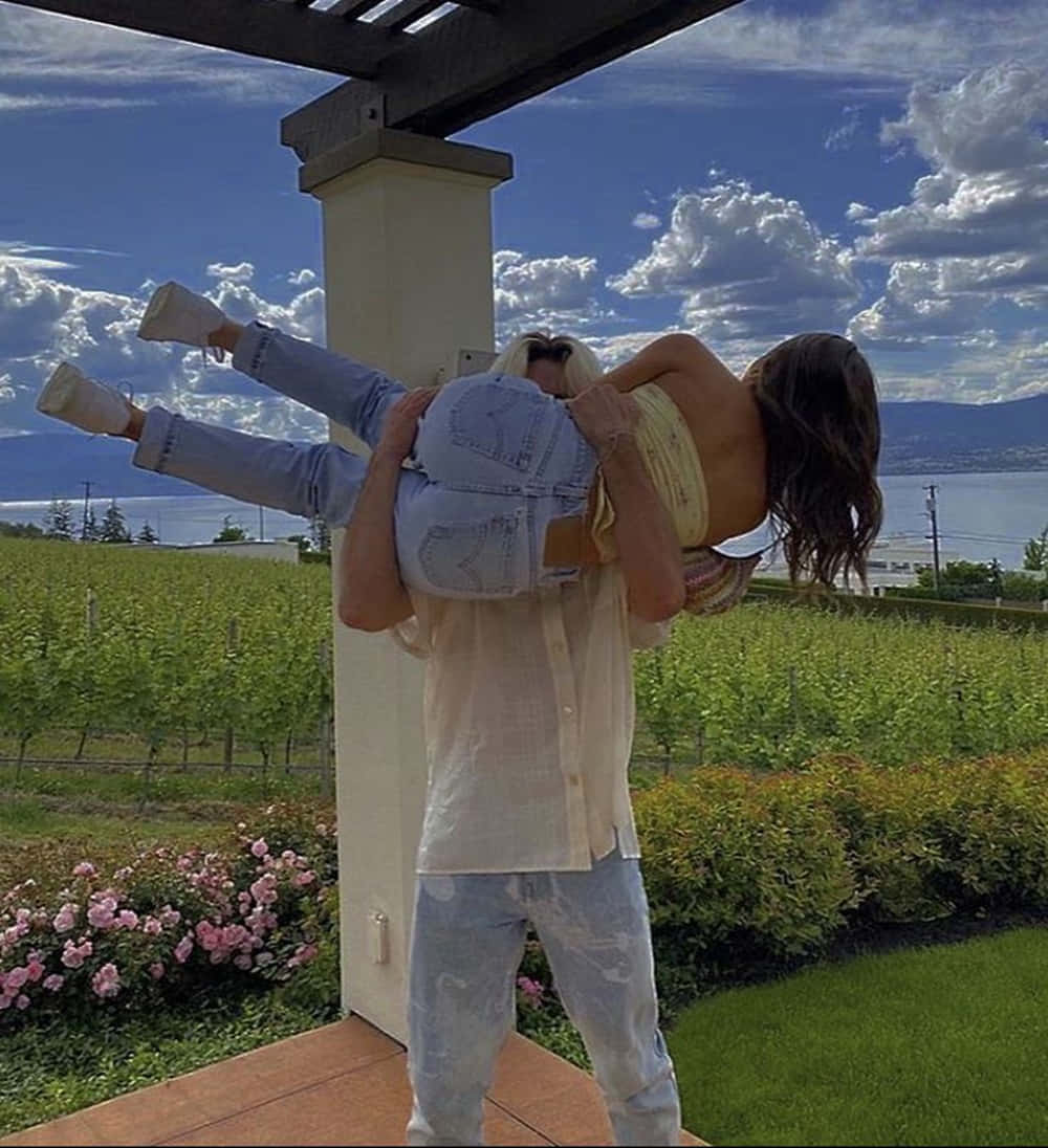 情侶藍天擁抱美照 壁紙 — 一個男人在藤架下背著一個女人，藤架下有葡萄園和湖泊，背景是明亮的藍天和蓬鬆的白雲。