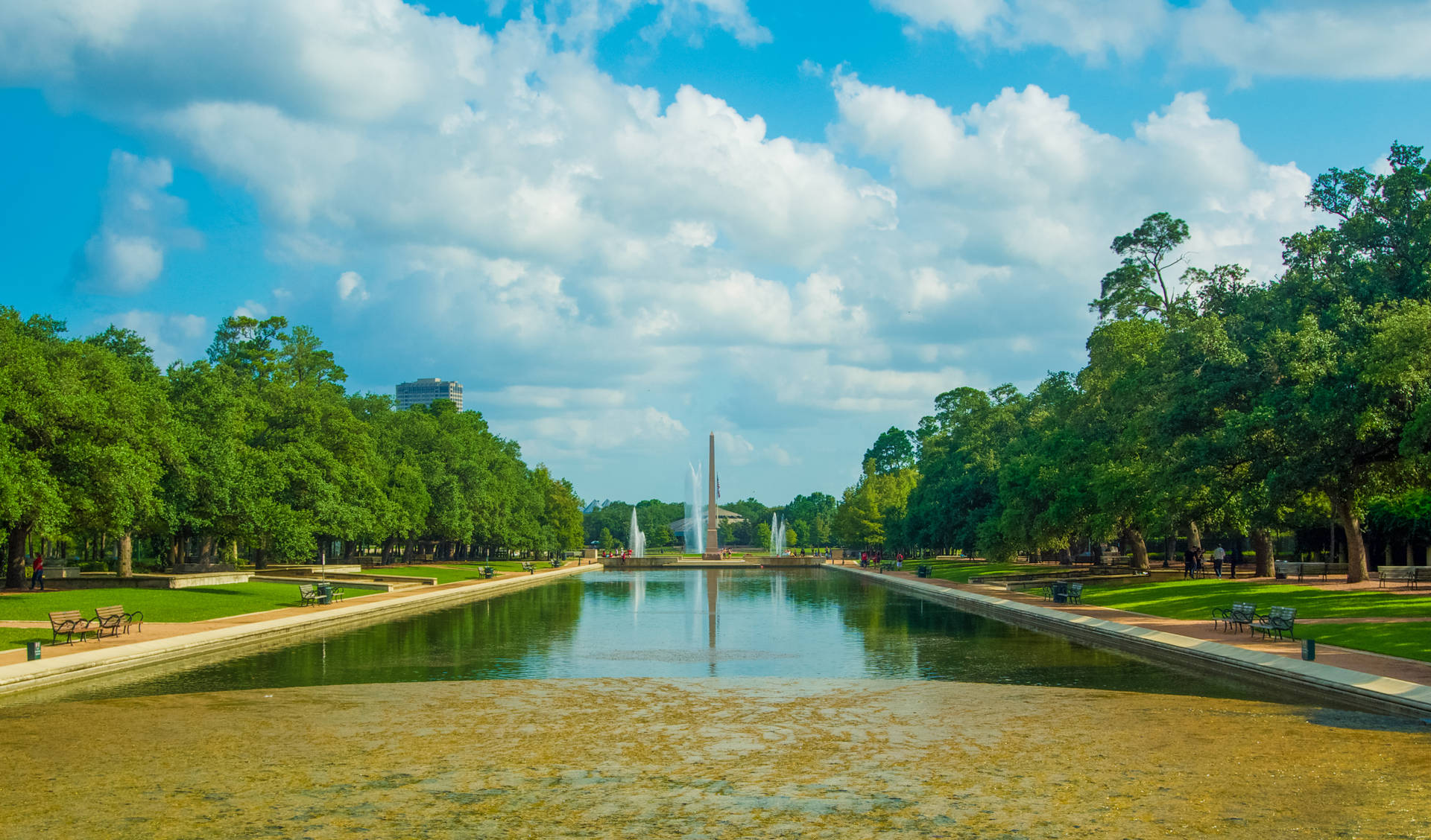 陽光明媚的天空下，公園運河寧靜，有噴泉和樹木。
