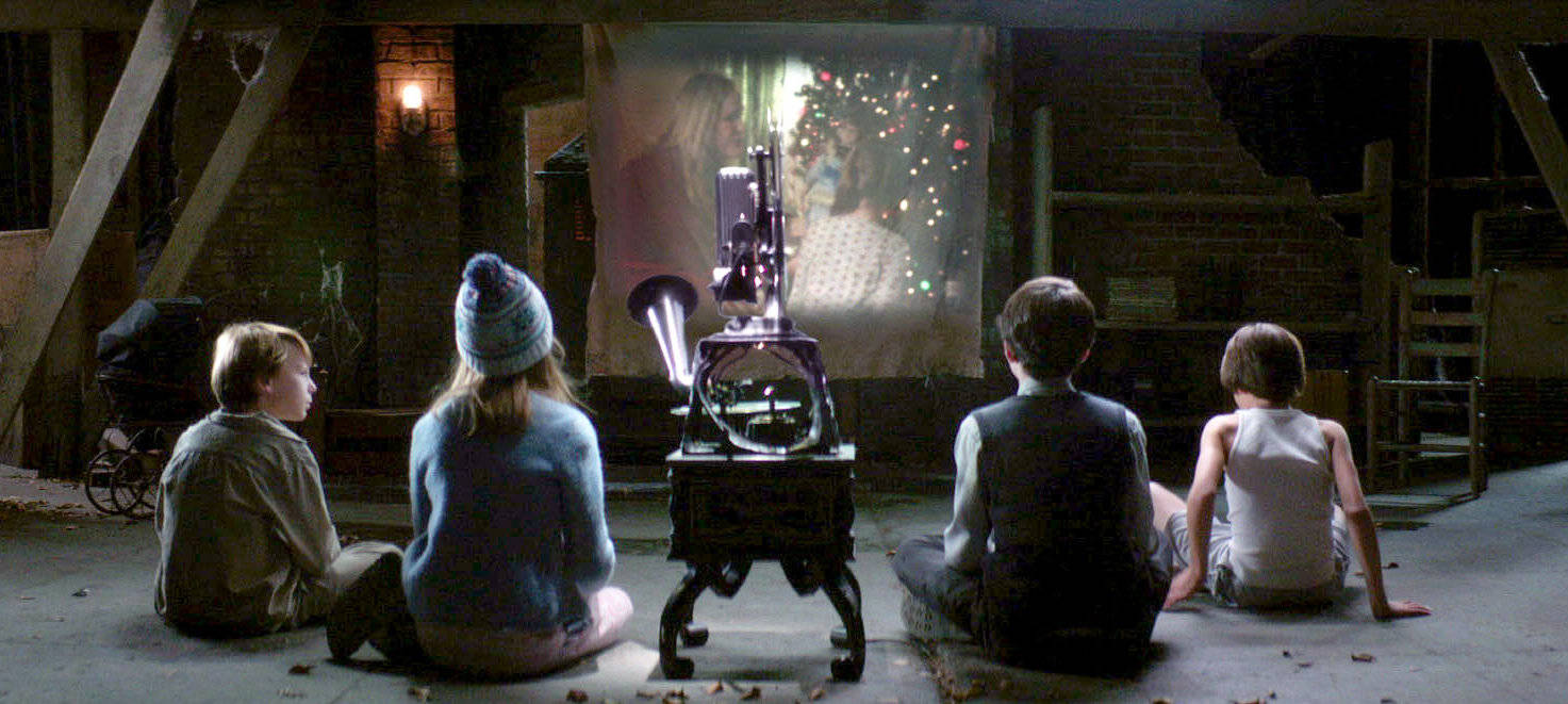 《惡靈古堡2》截圖 壁紙 — 四個孩子盤腿坐在混凝土地板上，看著一部復古投影機將發光的影片投射到鄉村穀倉的磚牆上。