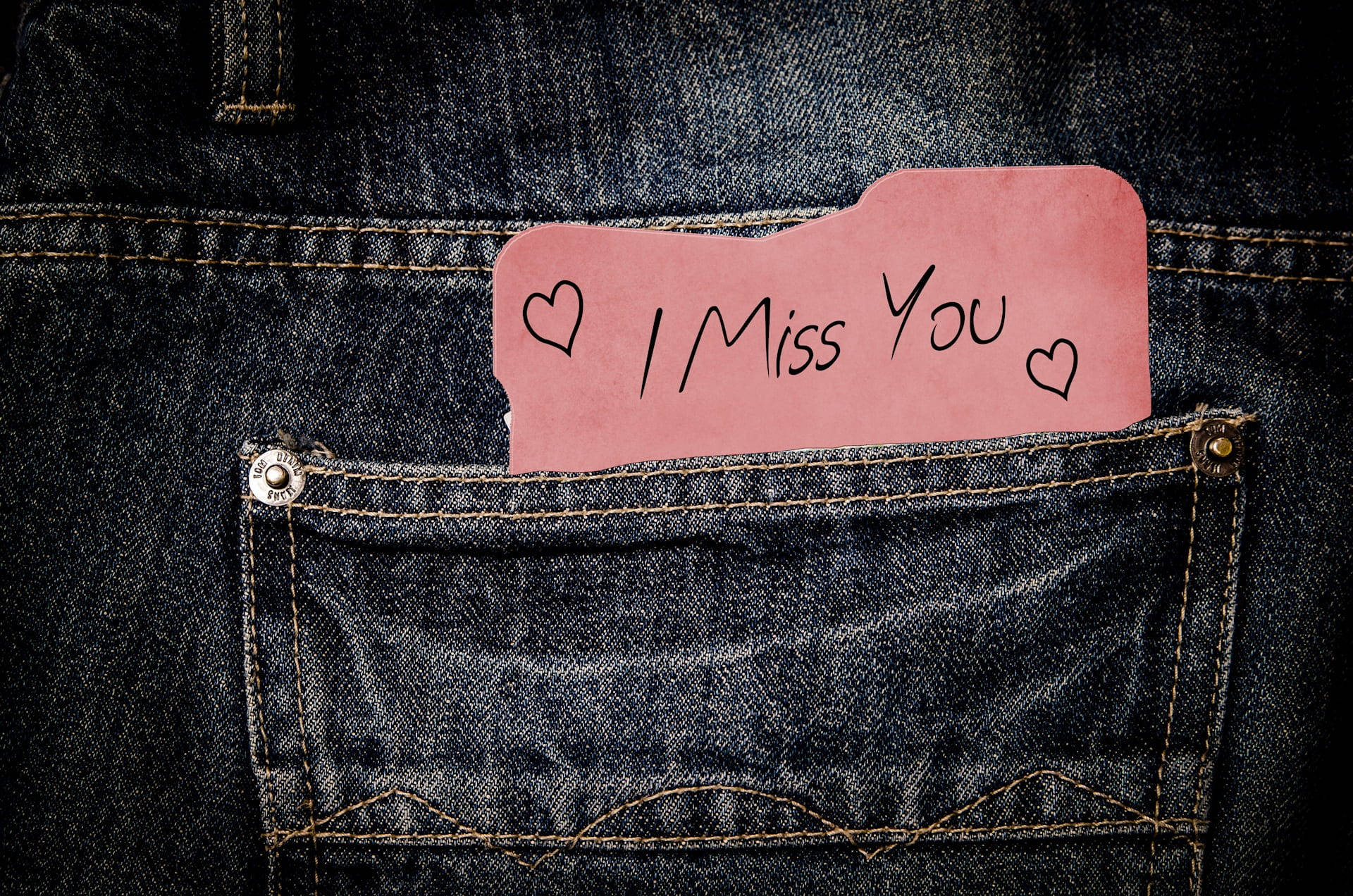 想念你 牛仔口袋便條 壁紙 — 一張粉紅色手寫便條，上面寫著「I Miss You」並帶有心形圖案，塞在深色牛仔褲的後袋裡。
