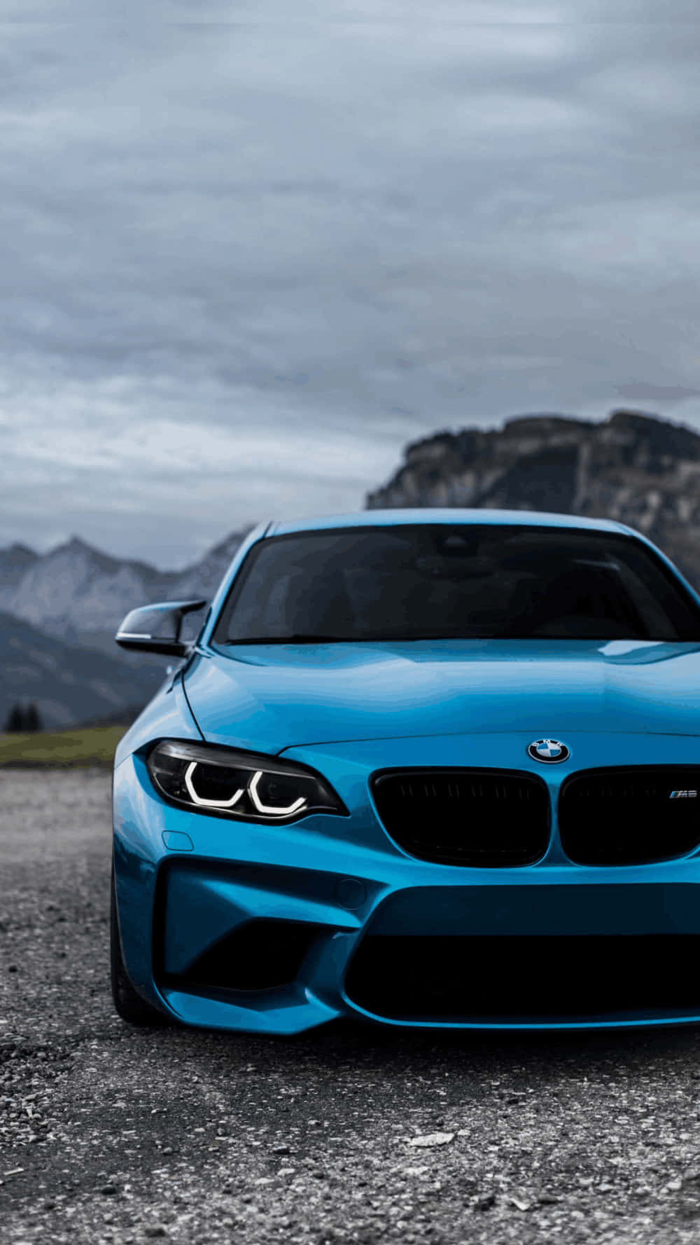 藍色BMW M2跑車停泊在碎石上，靠近山脈，天空多雲。