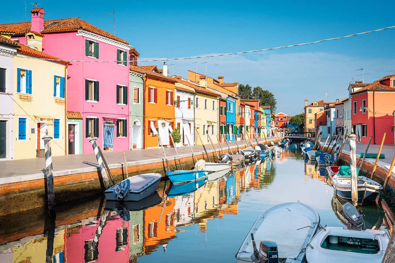 Italian Island 桌布 壁紙 — 意大利布拉諾島寧靜的運河兩旁排列著色彩繽紛的粉彩房屋，船隻停泊，清澈的藍天倒映在水中。