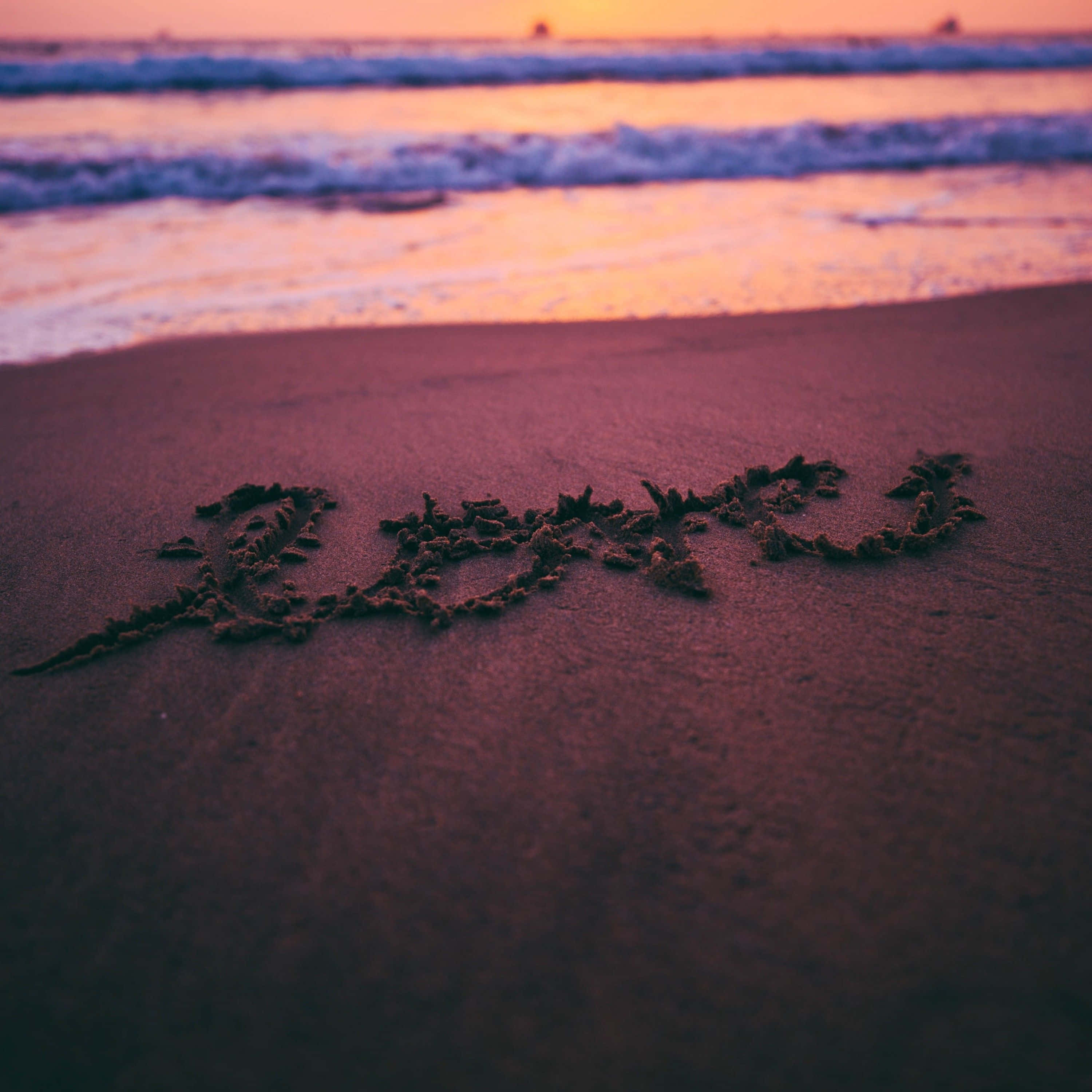 Beach Love 桌布 壁紙 — 「愛」這個字寫在日落時分海灘的沙灘上，溫暖的橙色和紫色色調倒映在水面上。